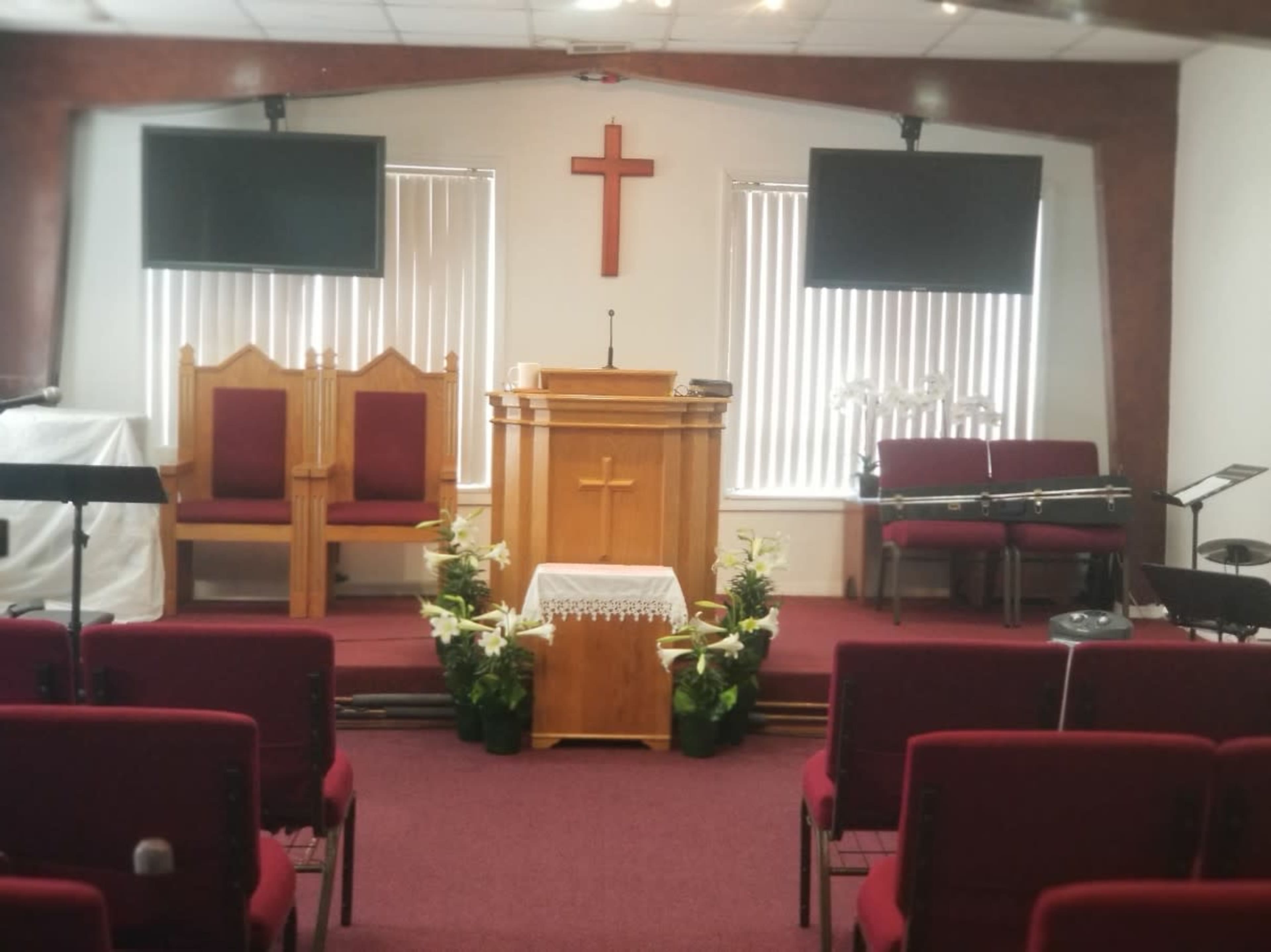 The image shows the interior of a church, featuring a wooden pulpit, two wooden chairs, and seating arranged in rows with a cross mounted on the wall above the pulpit.