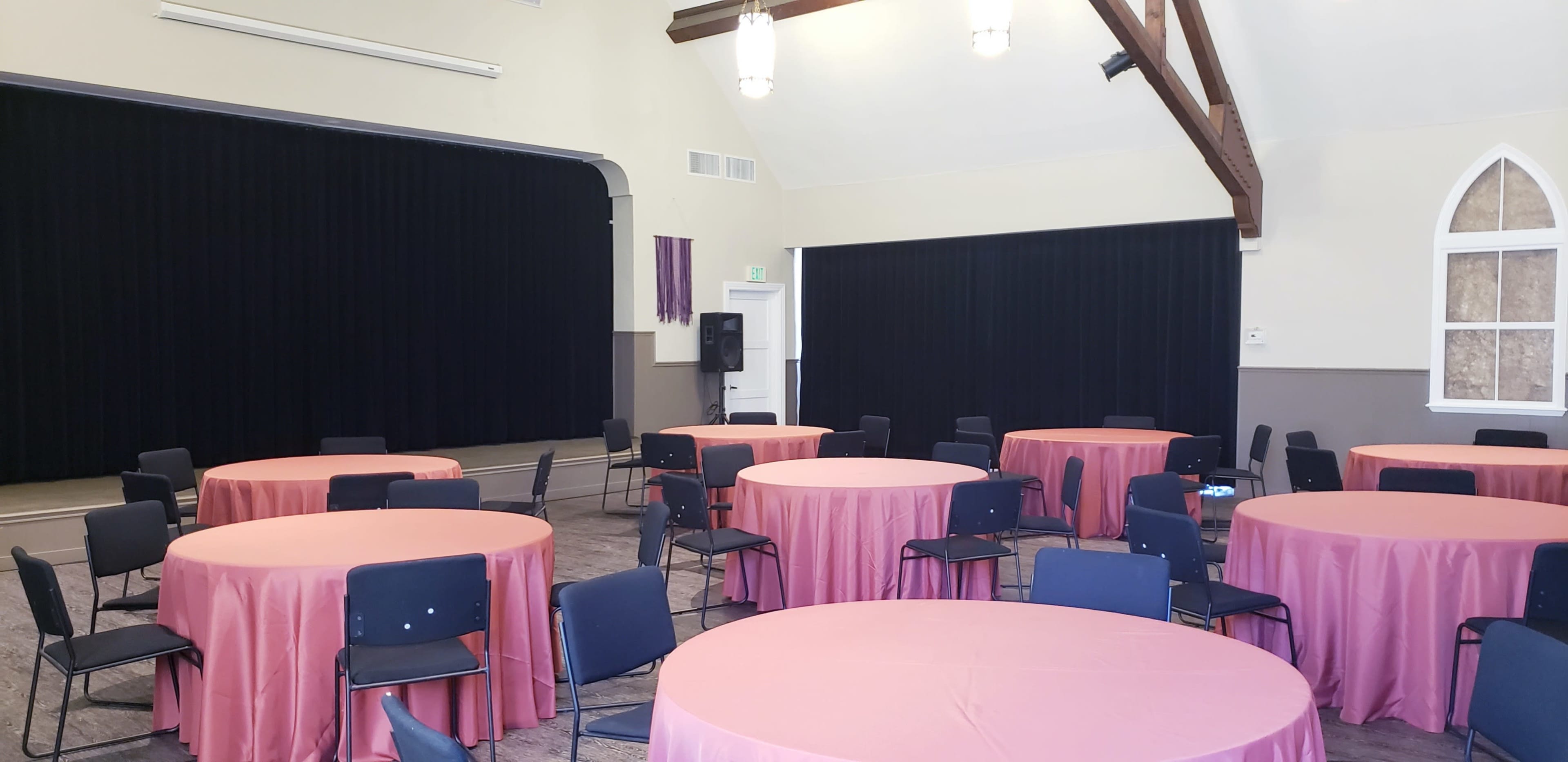A spacious room features several round tables covered with pink tablecloths, arranged with black chairs, and a stage area with black curtains at one end.