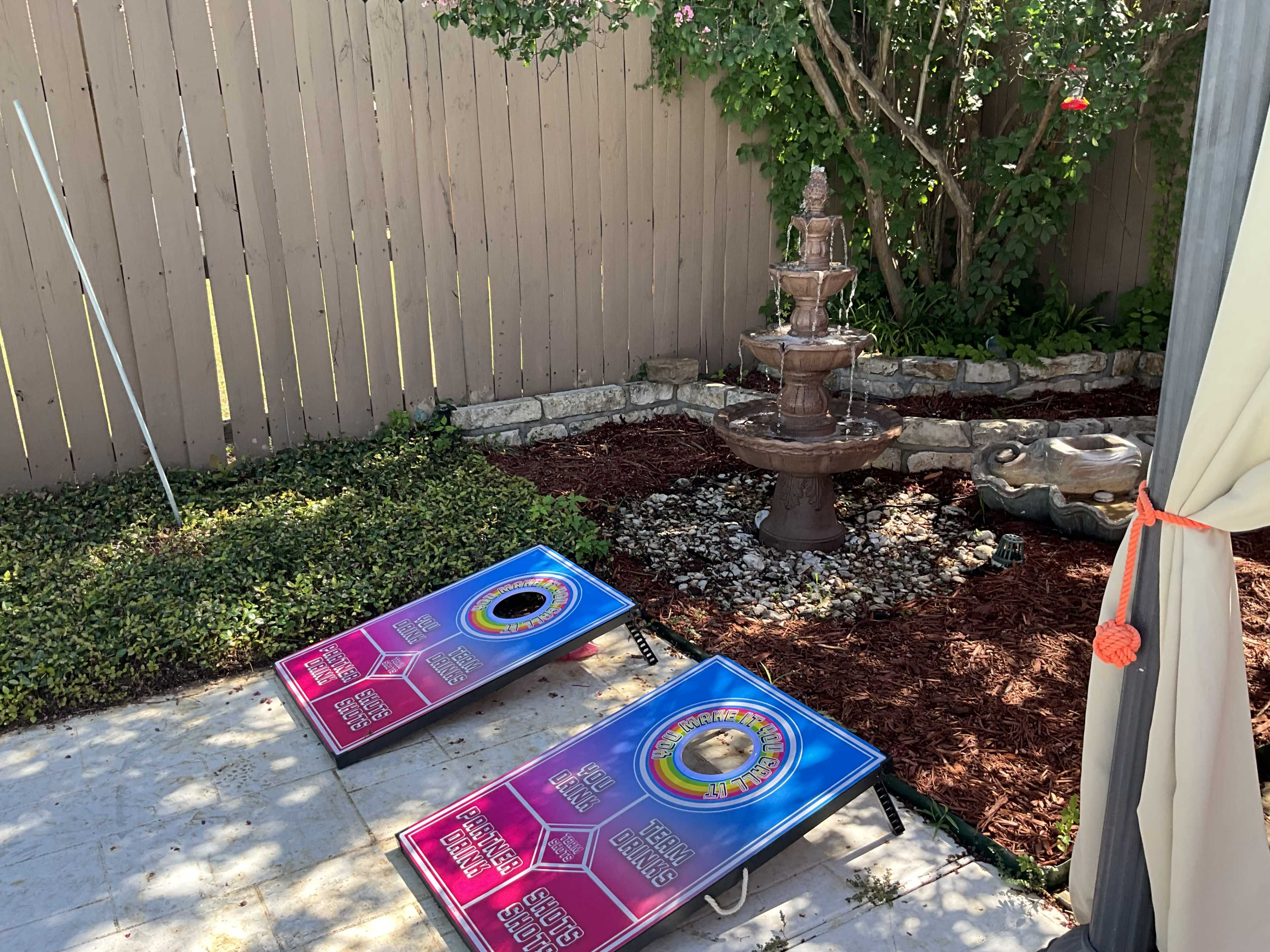 A backyard area with a fountain surrounded by rocks and plants, along with two colorful cornhole boards positioned nearby.