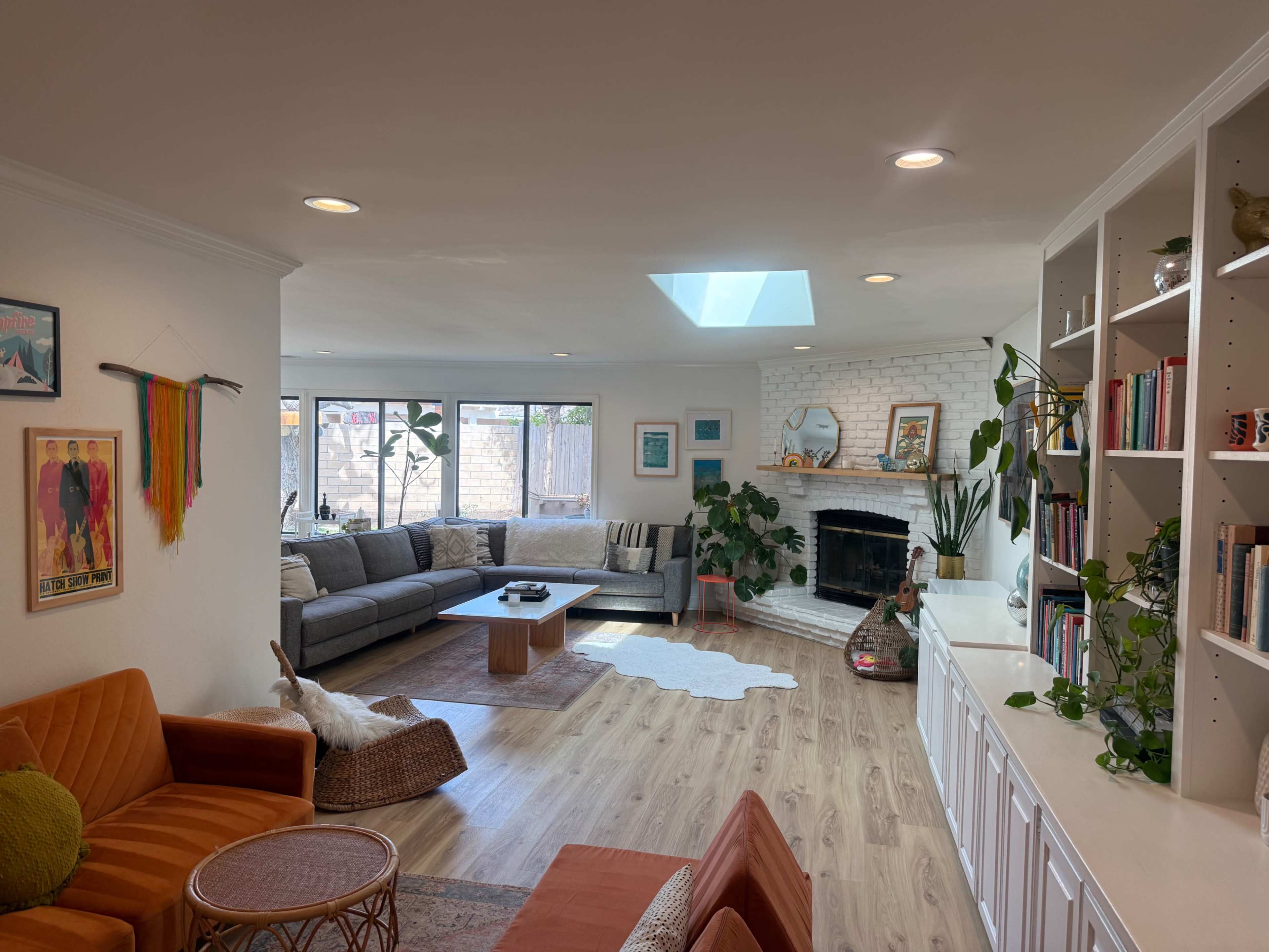 A bright living room features a gray sectional sofa, a wooden coffee table, and a white brick fireplace, with large windows letting in natural light and greenery in the decor.
