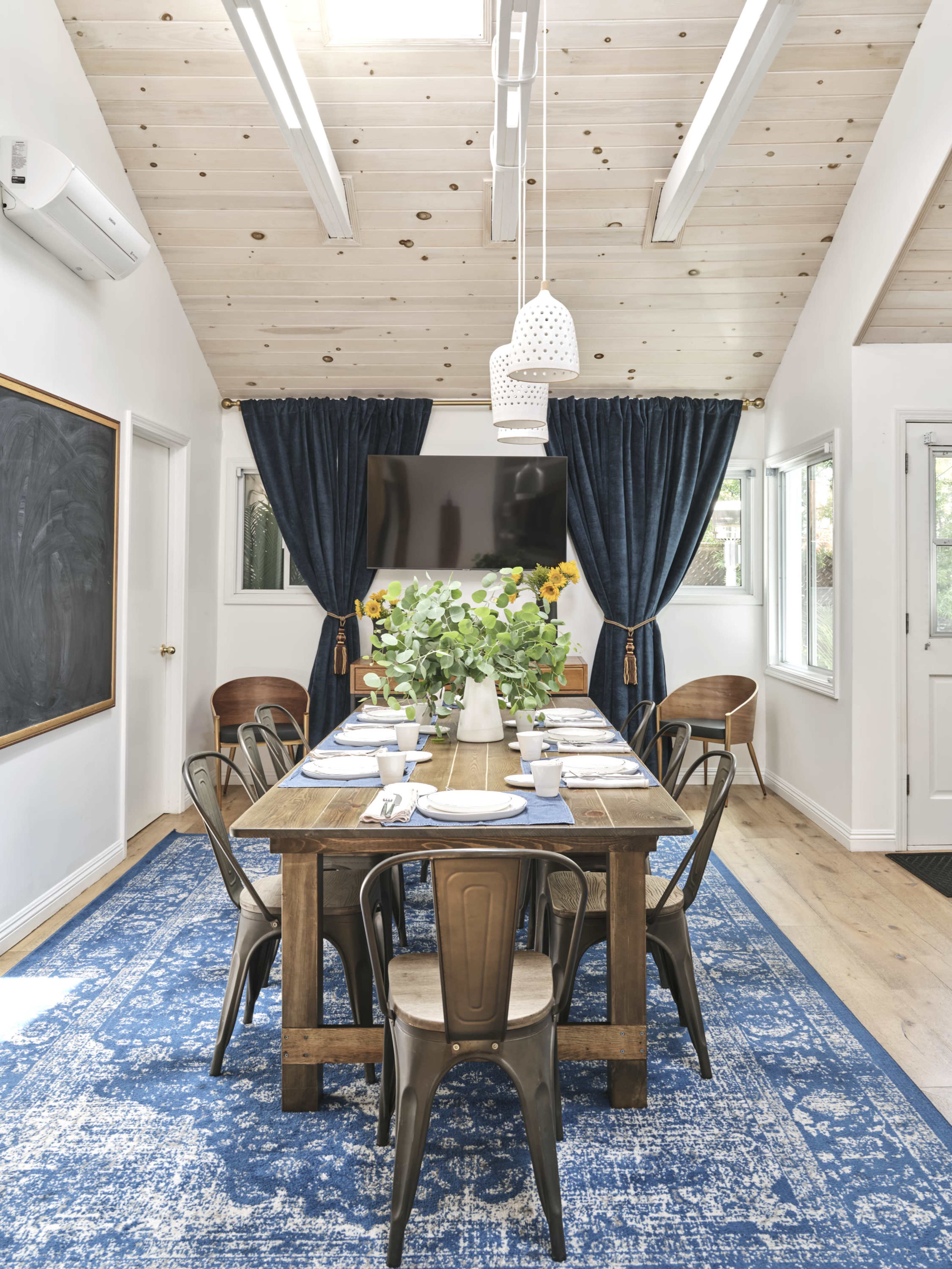 A dining room features a long wooden table set for a meal, surrounded by metal chairs, with blue curtains and a large television mounted on the wall.
