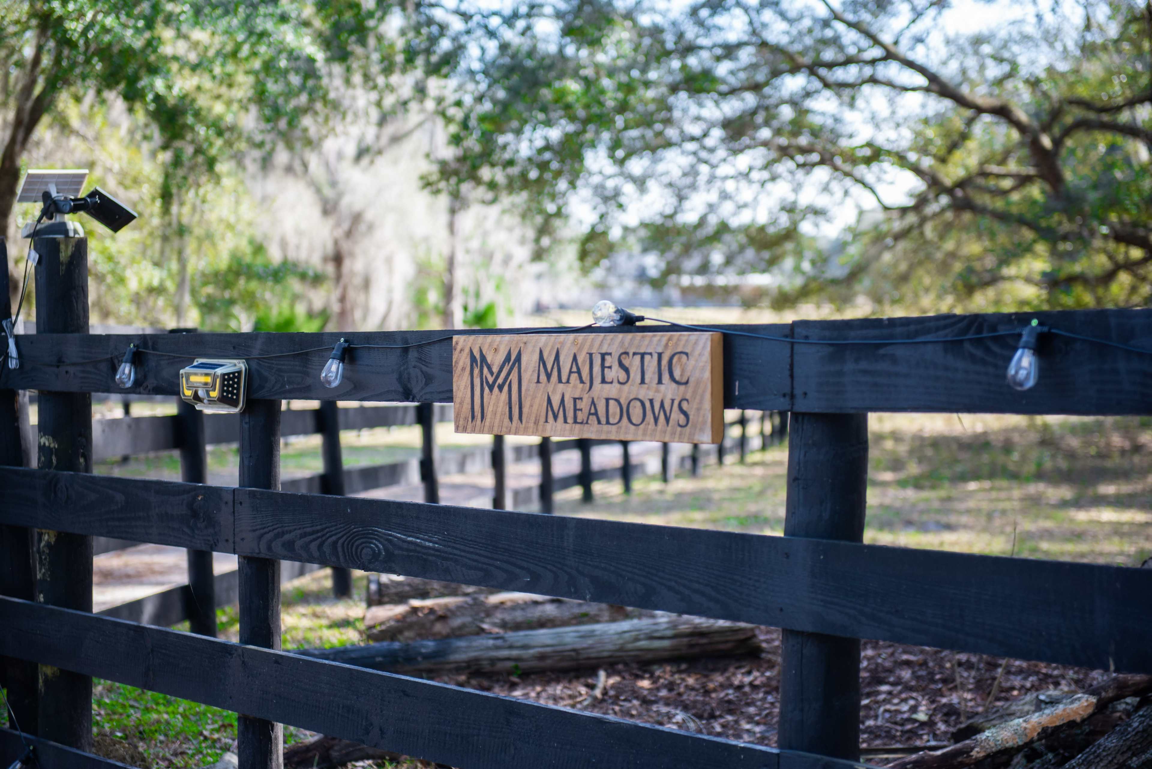A wooden sign reading "Majestic Meadows" hangs from a black wooden fence, surrounded by greenery and trees.