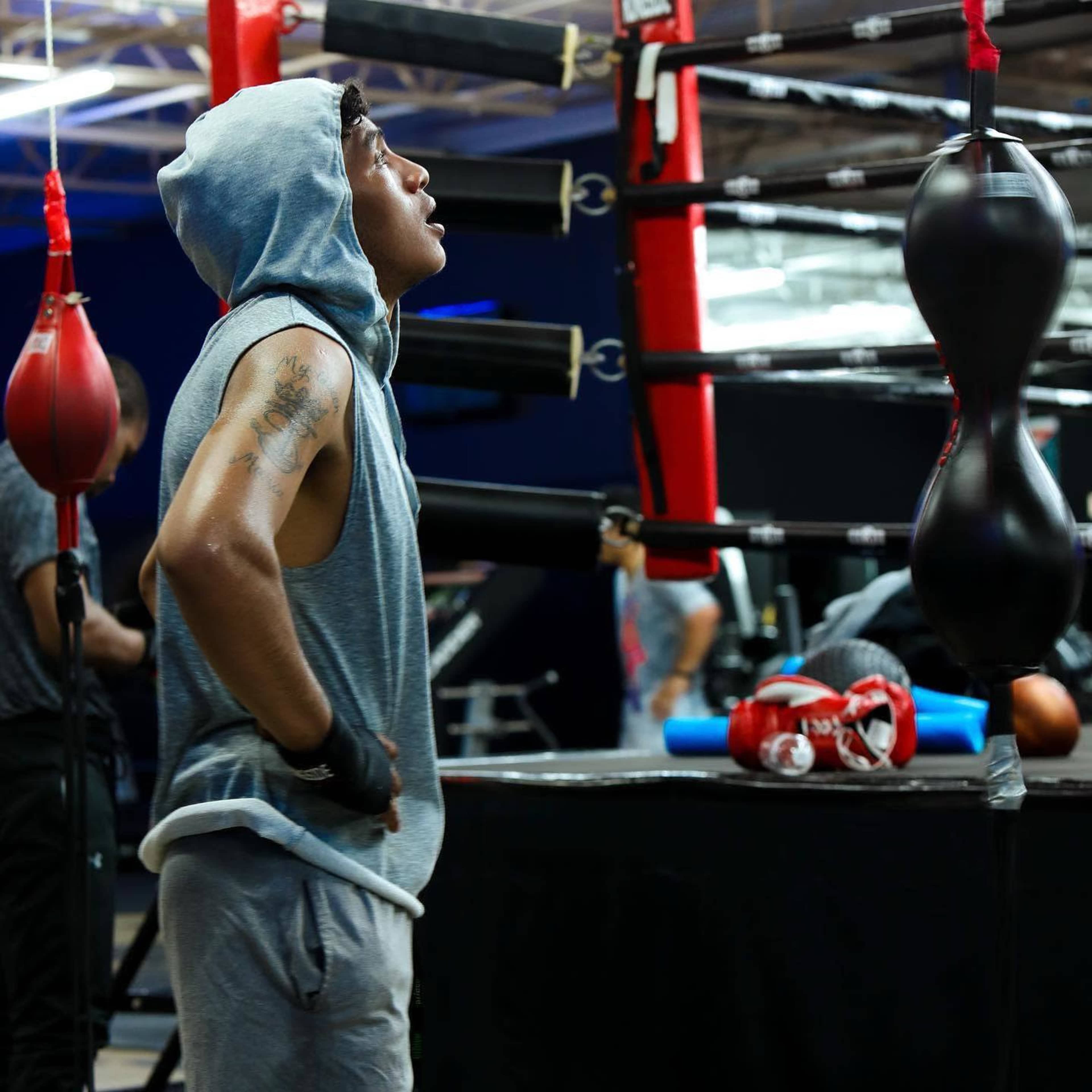 A boxer in a gray hoodie looks up at a speed bag in a gym surrounded by training equipment.