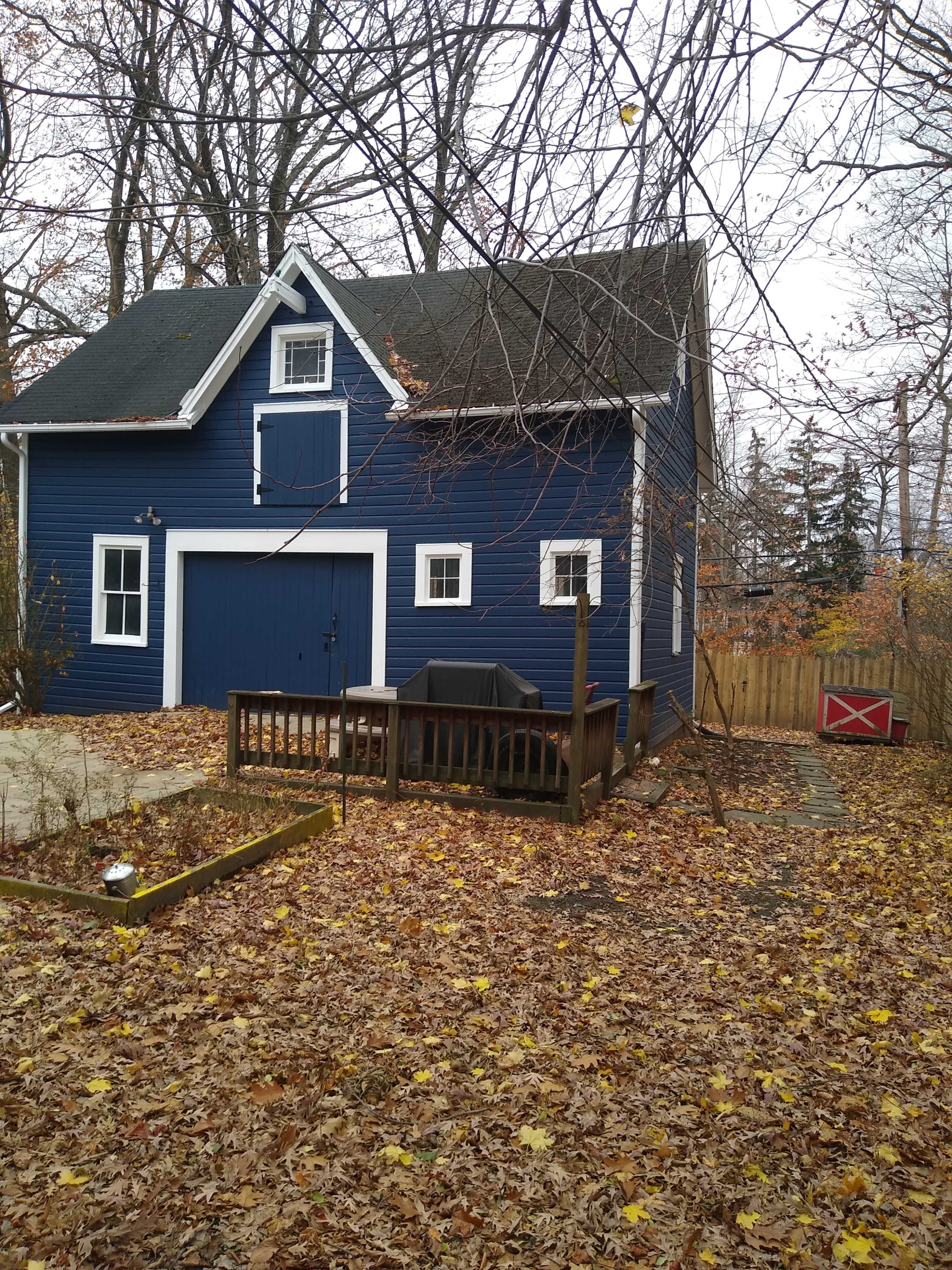 A blue two-story house with a deck, surrounded by fallen leaves and trees in a suburban setting.