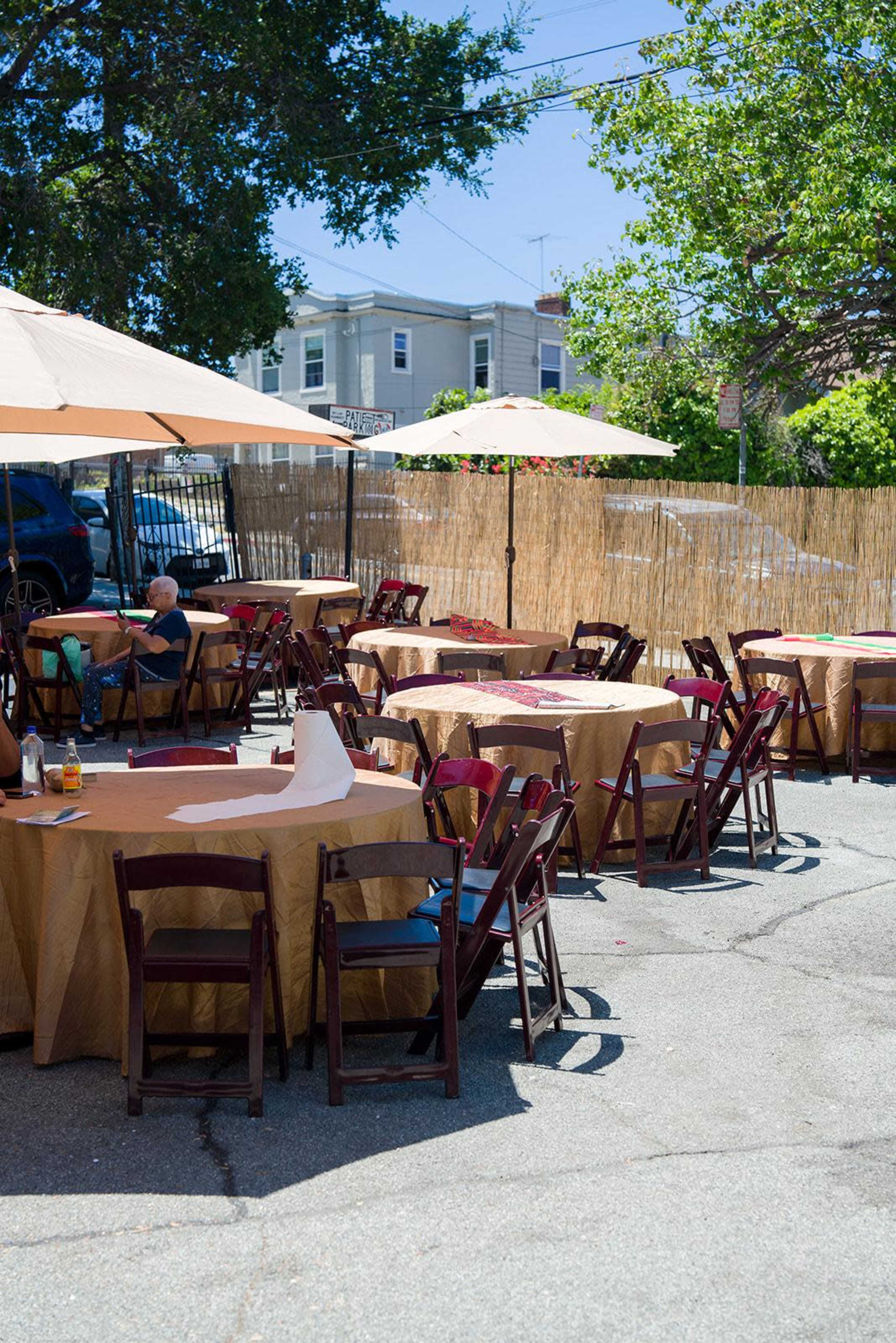 An outdoor dining area features several round tables with umbrellas, surrounded by chairs and set up on a paved surface.