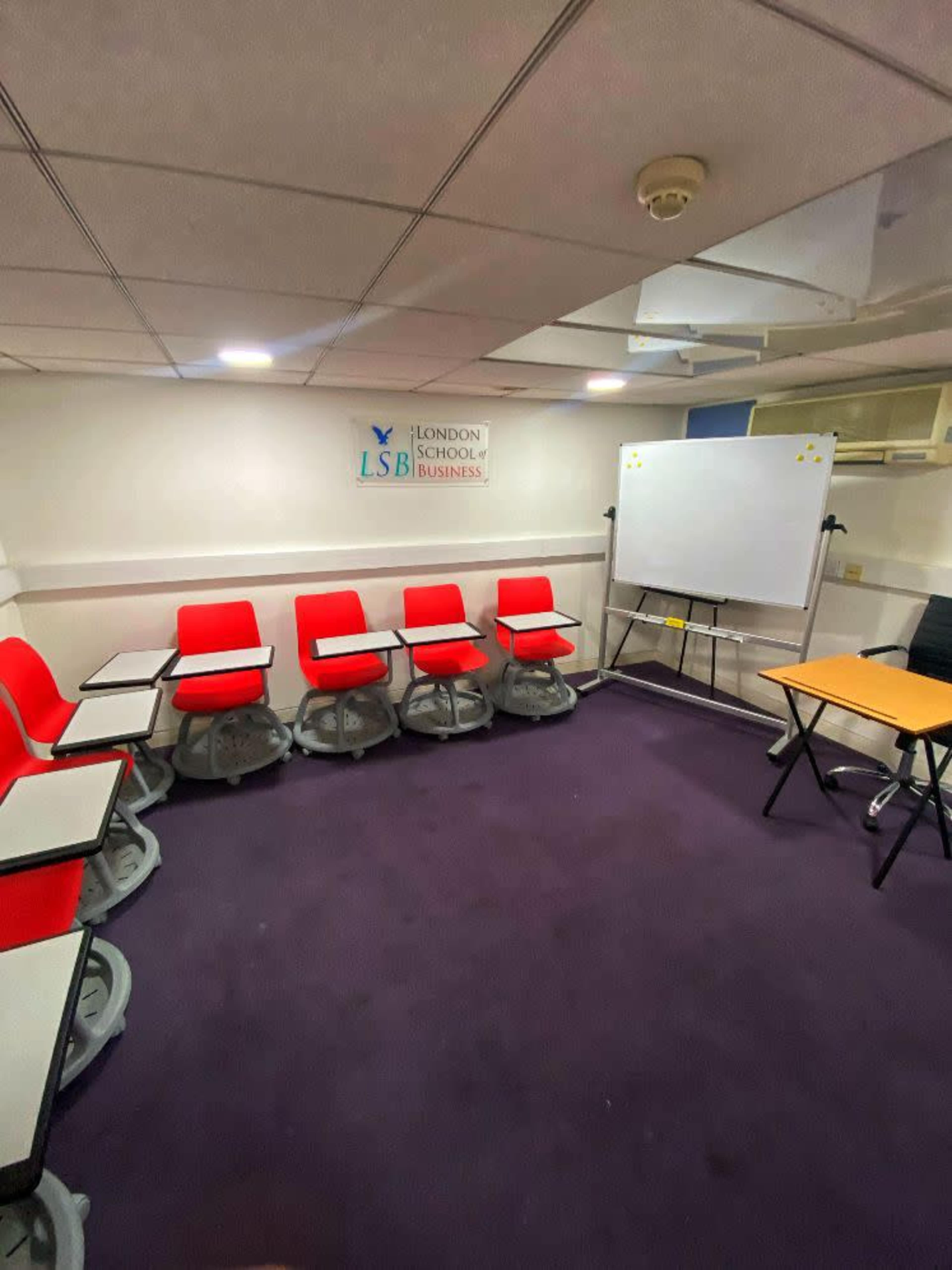The image shows a small classroom with red rolling chairs arranged in a semicircle around a whiteboard and a table against a purple carpeted floor.