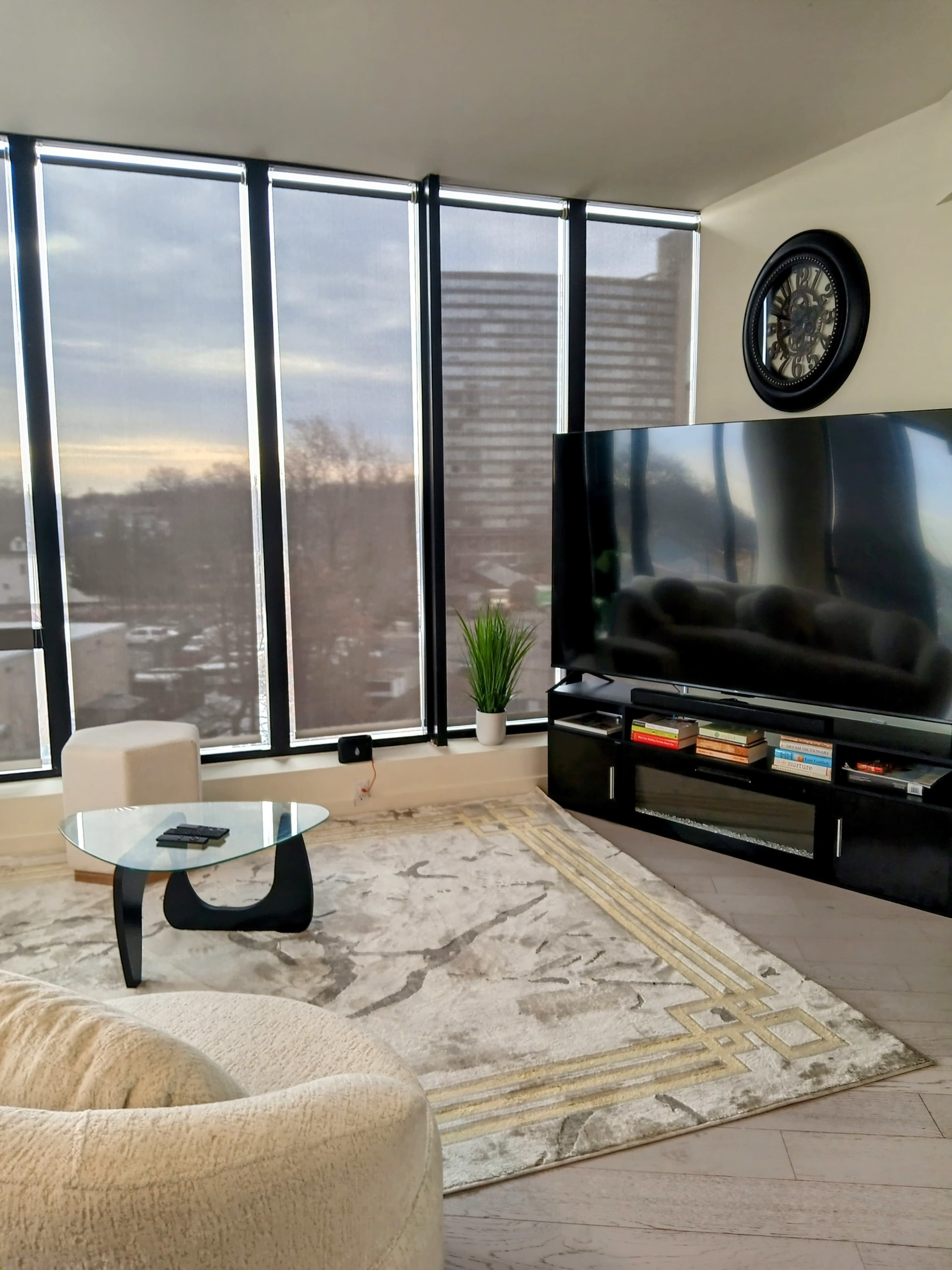 A living room features a large television, a round glass coffee table, and a cozy armchair, with floor-to-ceiling windows allowing natural light to fill the space.