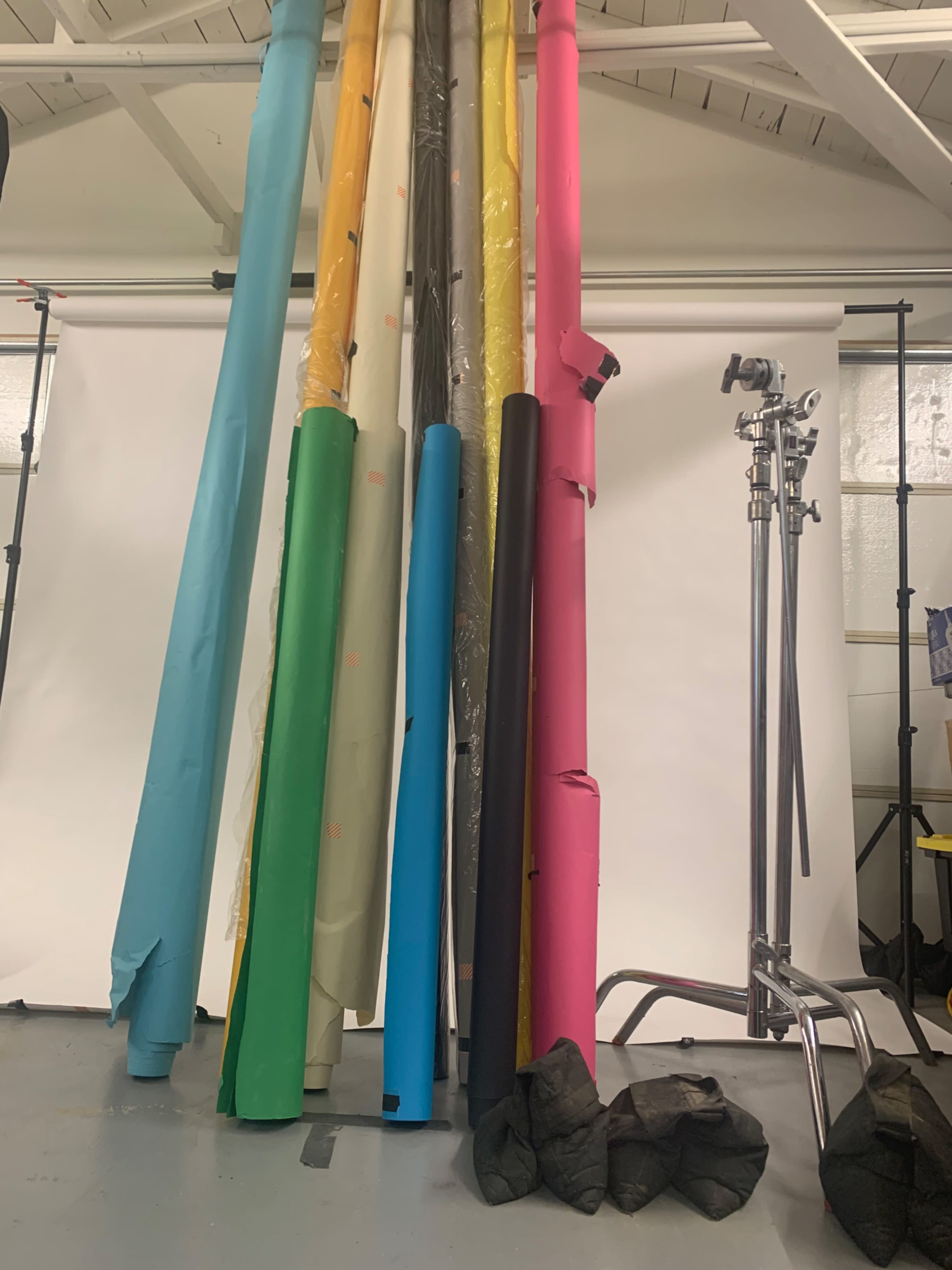 A collection of rolls of colored backdrop paper leaning against a wall in a studio, alongside a metal stand and black weights on the floor.