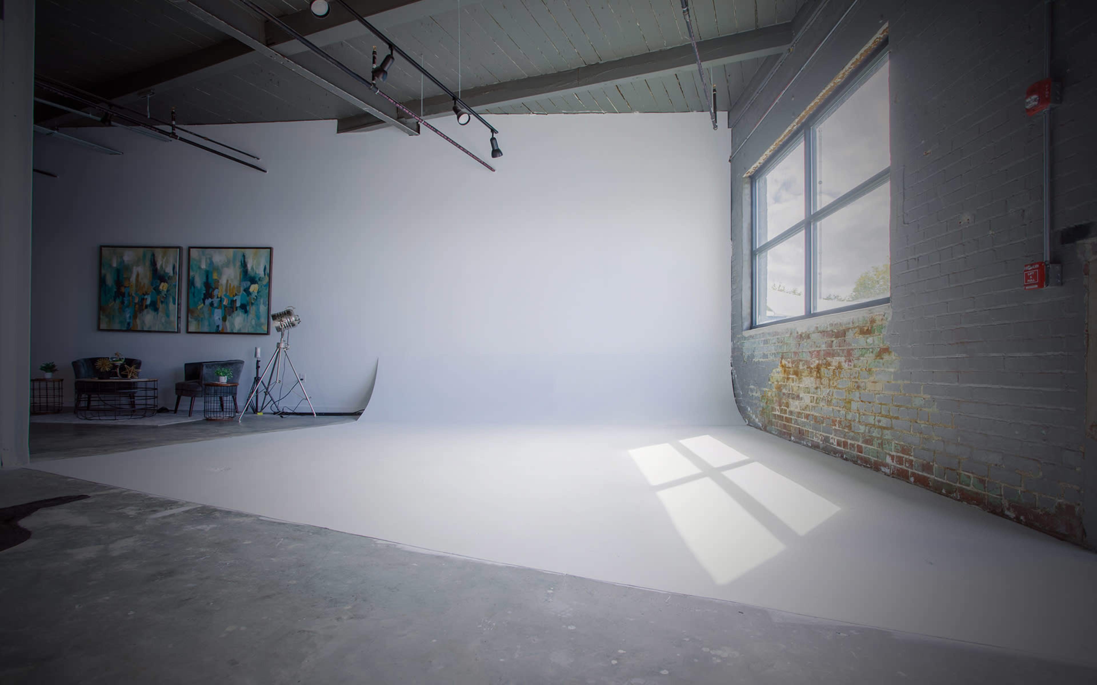 The image shows a bright, minimalist studio space with a white backdrop, large window, and two abstract paintings on the wall.