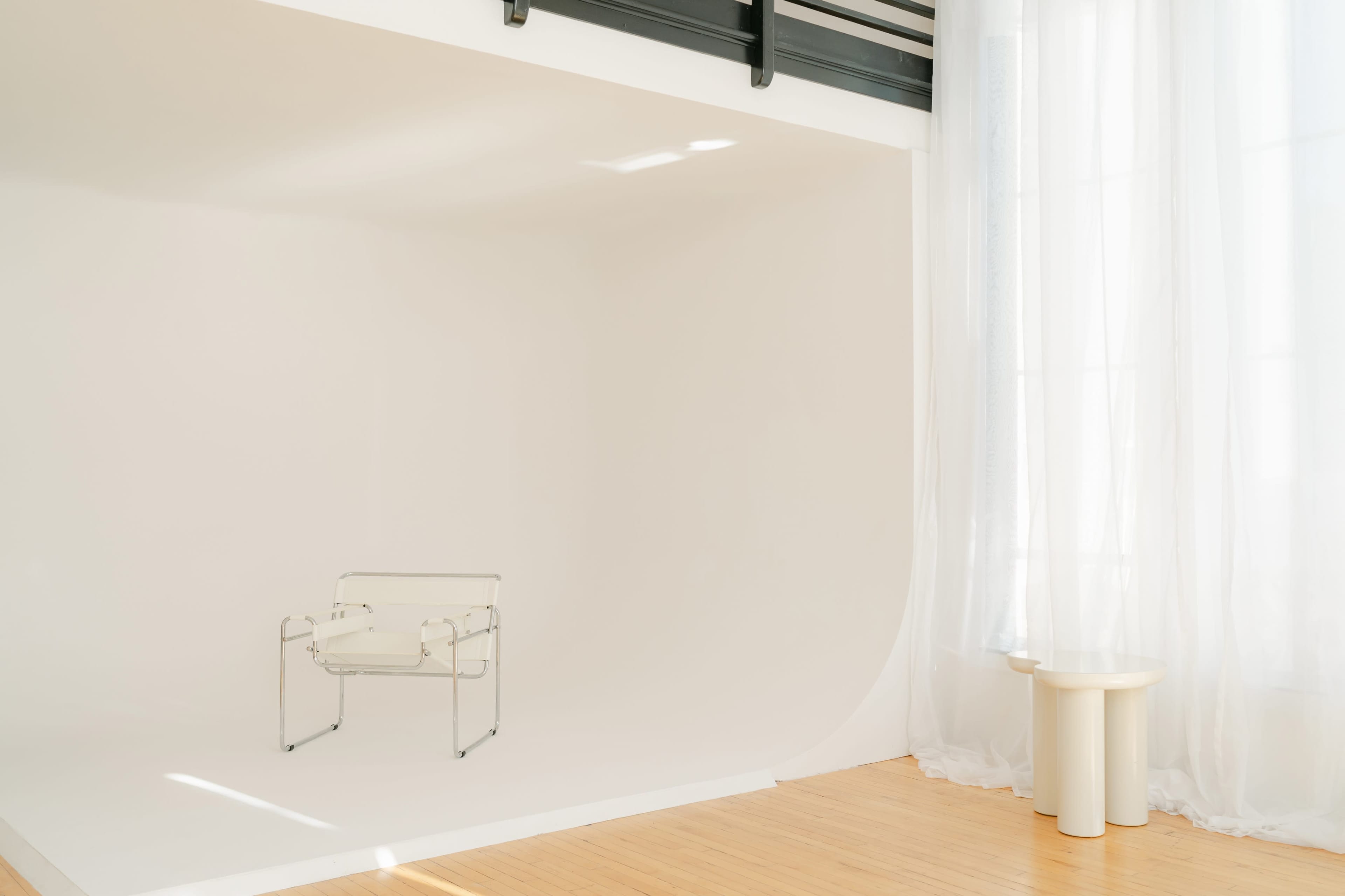 A clear chair sits in a minimalistic room with large windows and soft drapes, accompanied by a round pedestal table.