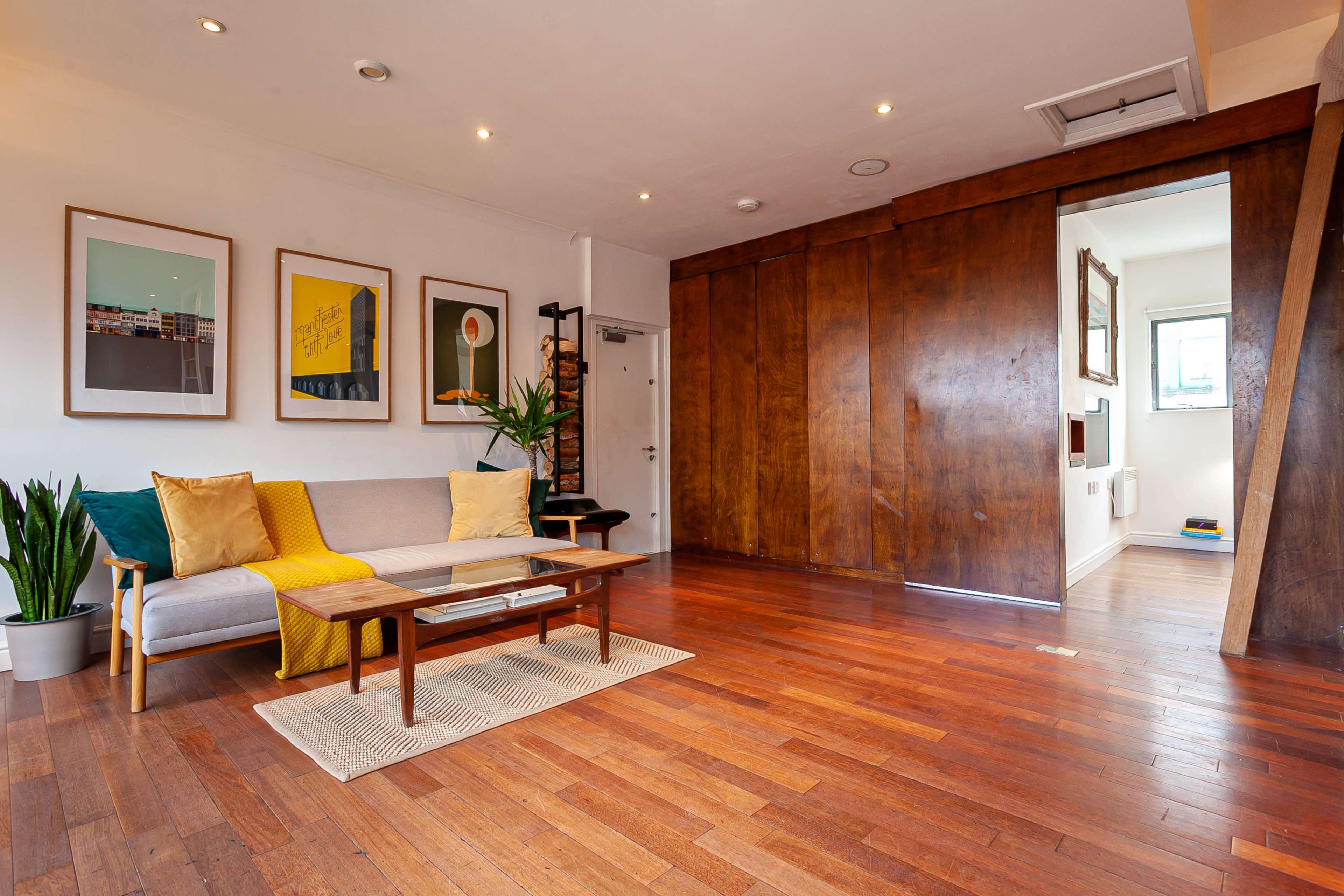 A living room features a sofa with yellow and gray cushions, a glass coffee table, framed artwork on the walls, and wooden flooring.
