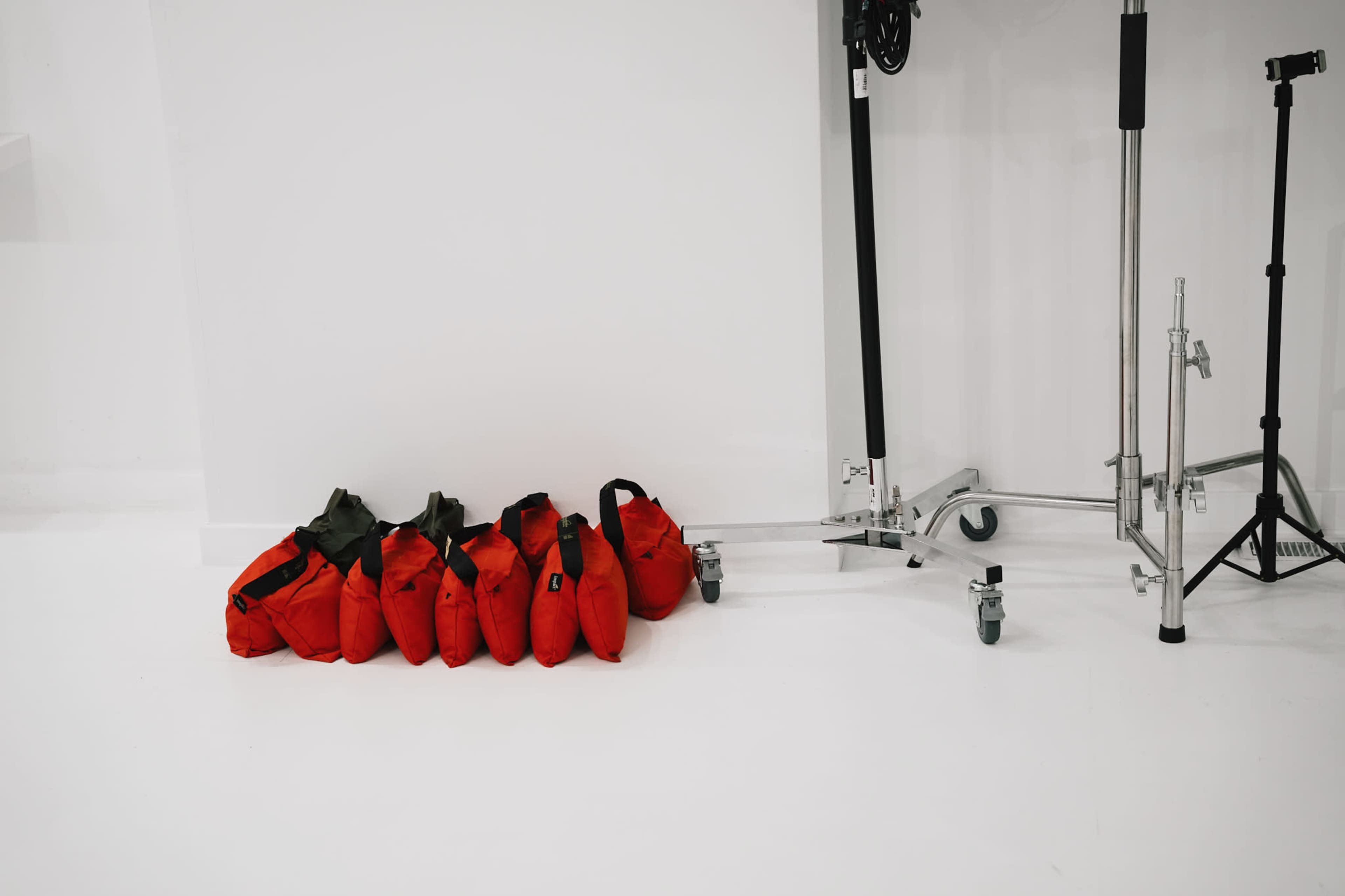 A row of six red bags is placed on a white floor next to a metal stand and a tripod.