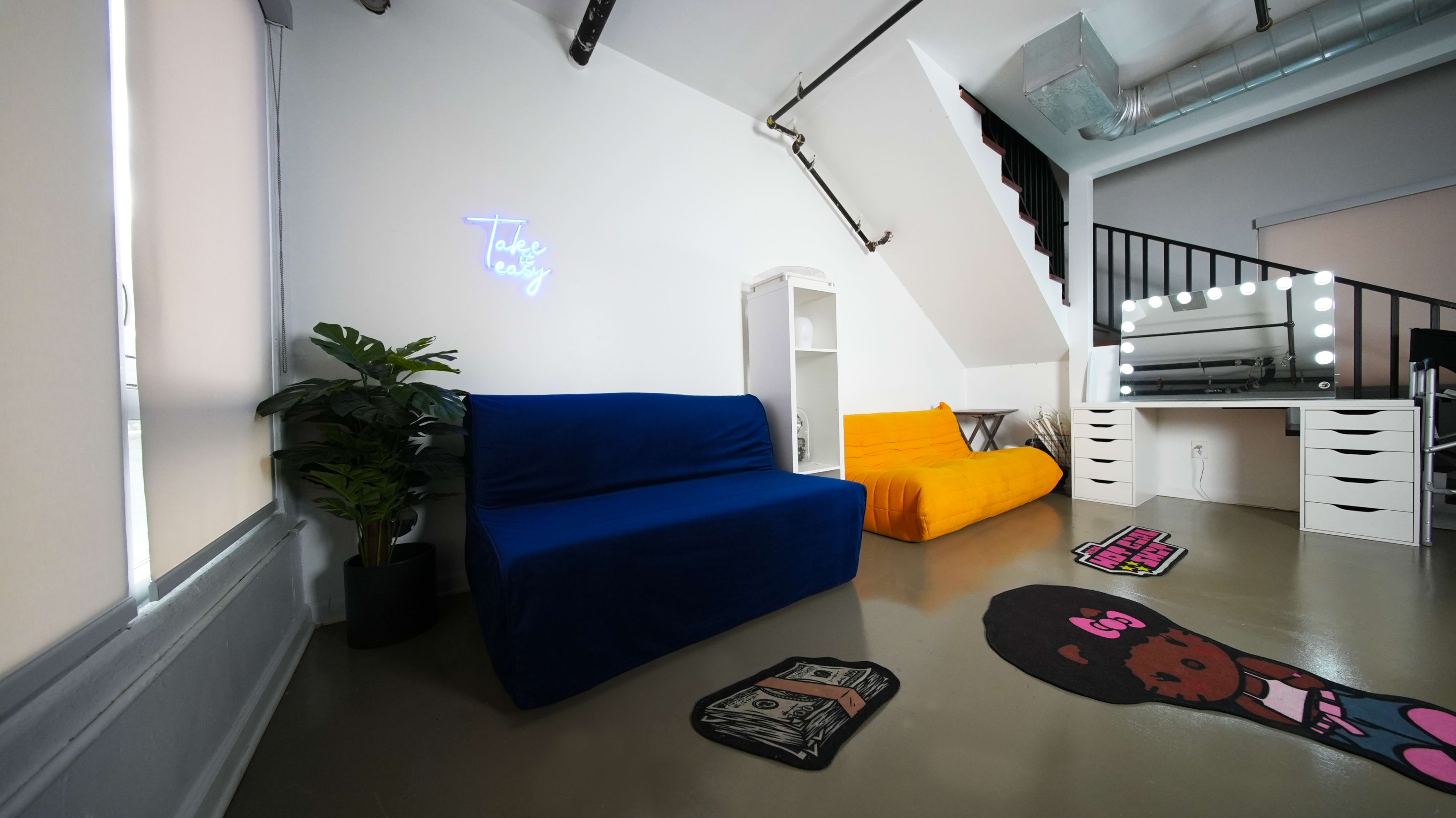 The image shows a modern interior space with a blue and yellow sofa, a potted plant, a mirrored vanity with lights, and a staircase in the background.
