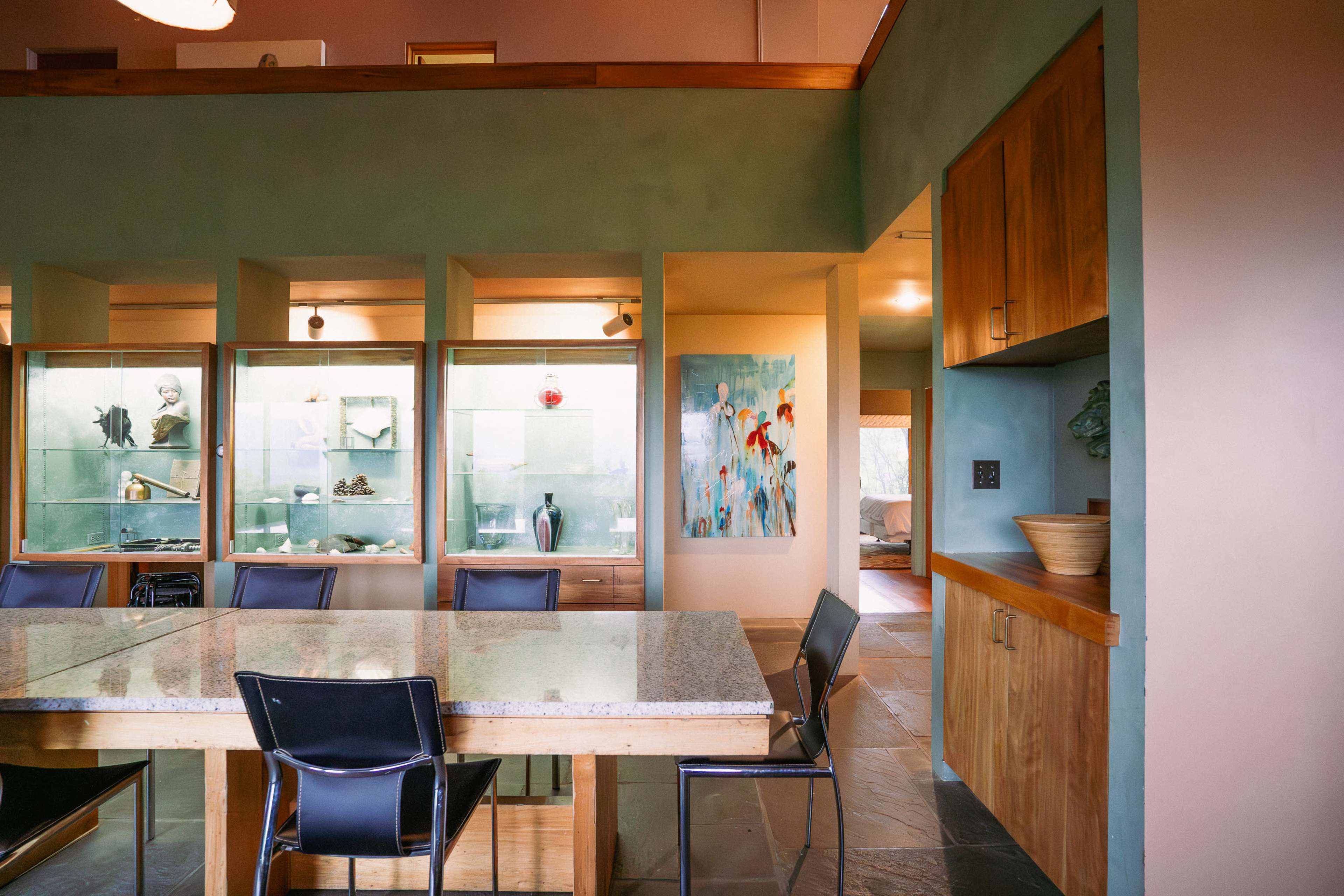 A modern dining area features a granite table surrounded by black chairs, with display cabinets showcasing decorative items and a colorful painting on the wall.