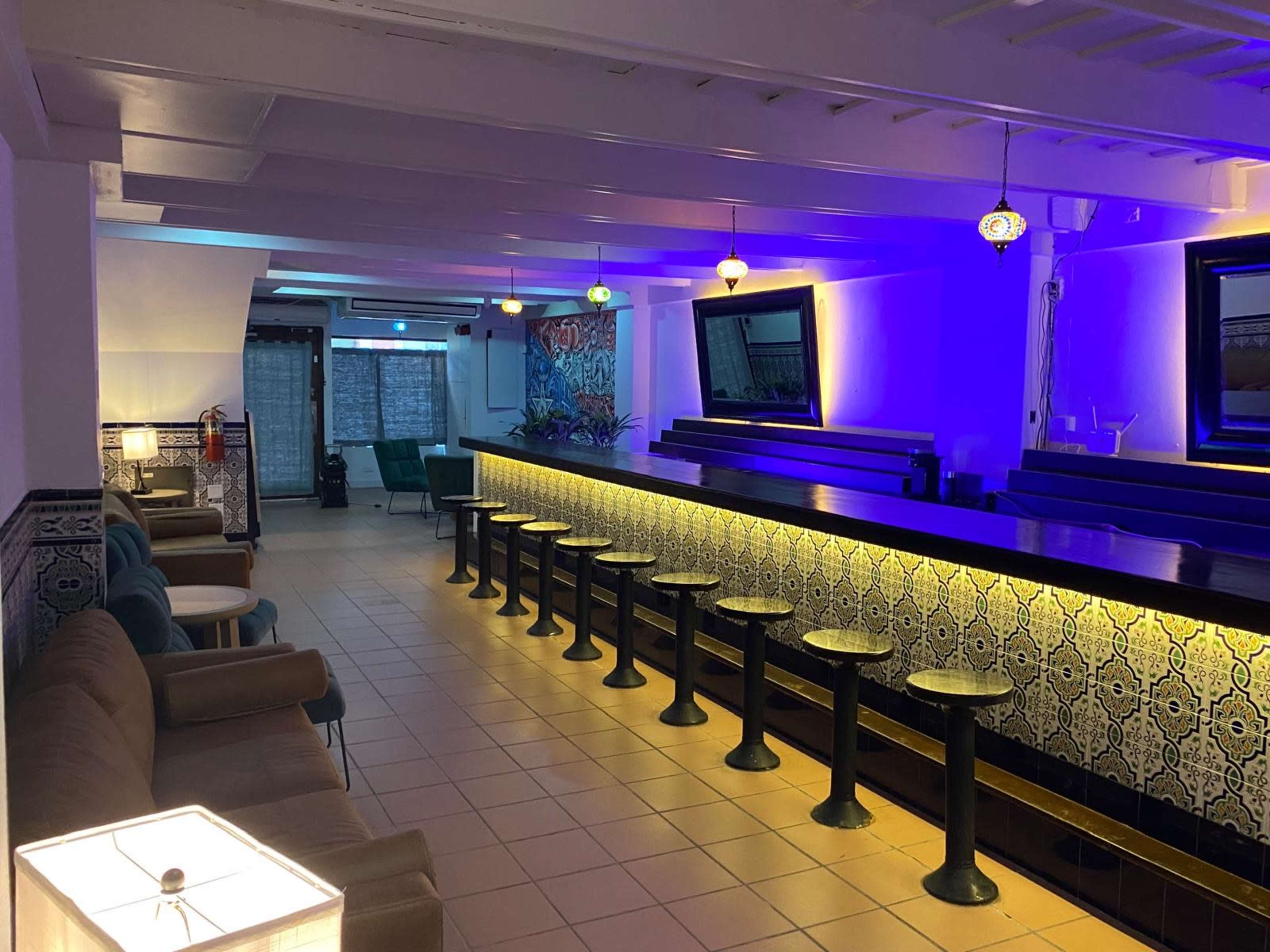 The image depicts a modern, well-lit lounge area featuring a long bar with stools, seating along one wall, and decorative lights illuminating the space.