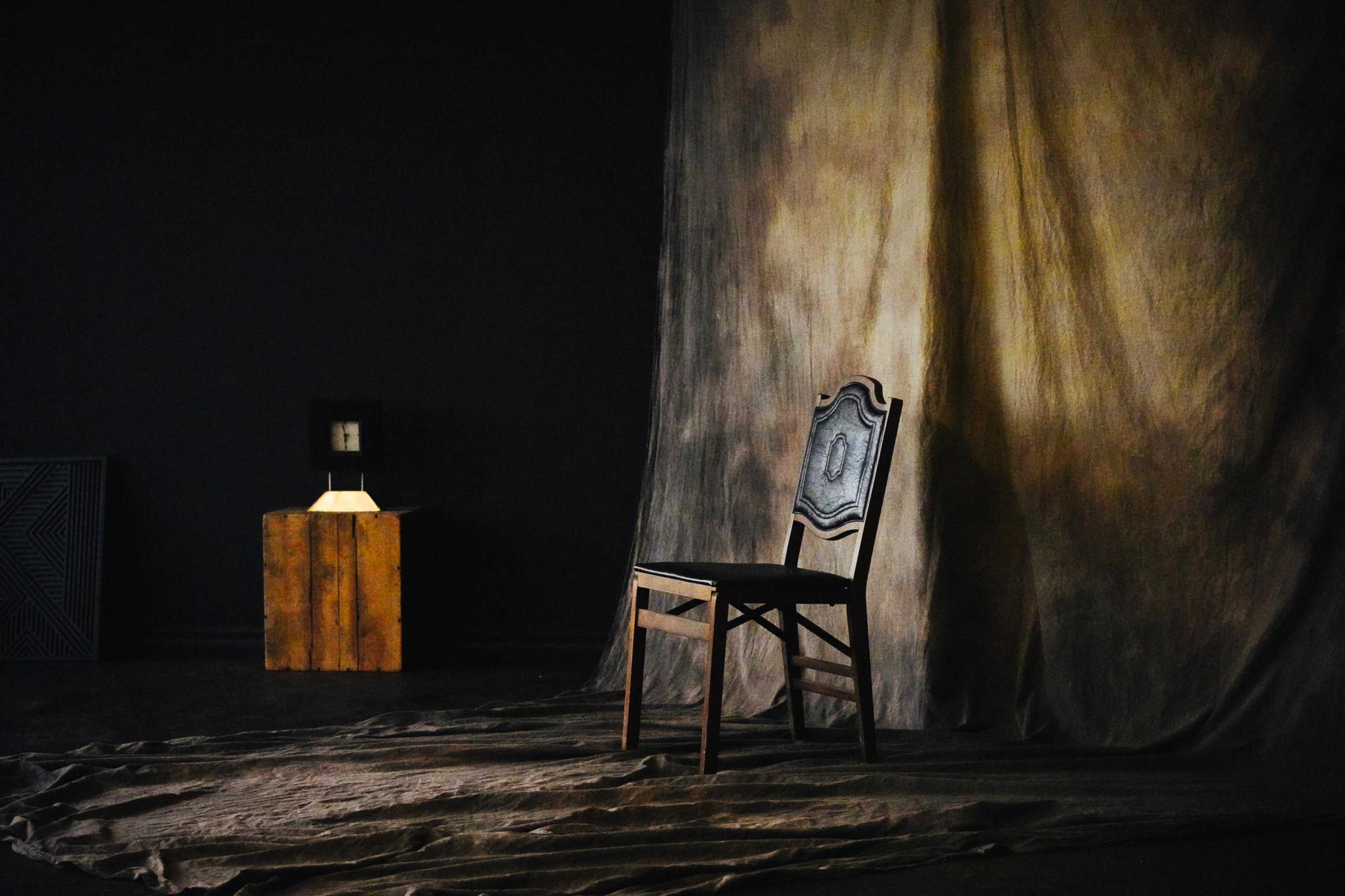 The image features a wooden chair positioned on a draped fabric floor, accompanied by a small table with a lamp in a dimly lit room.