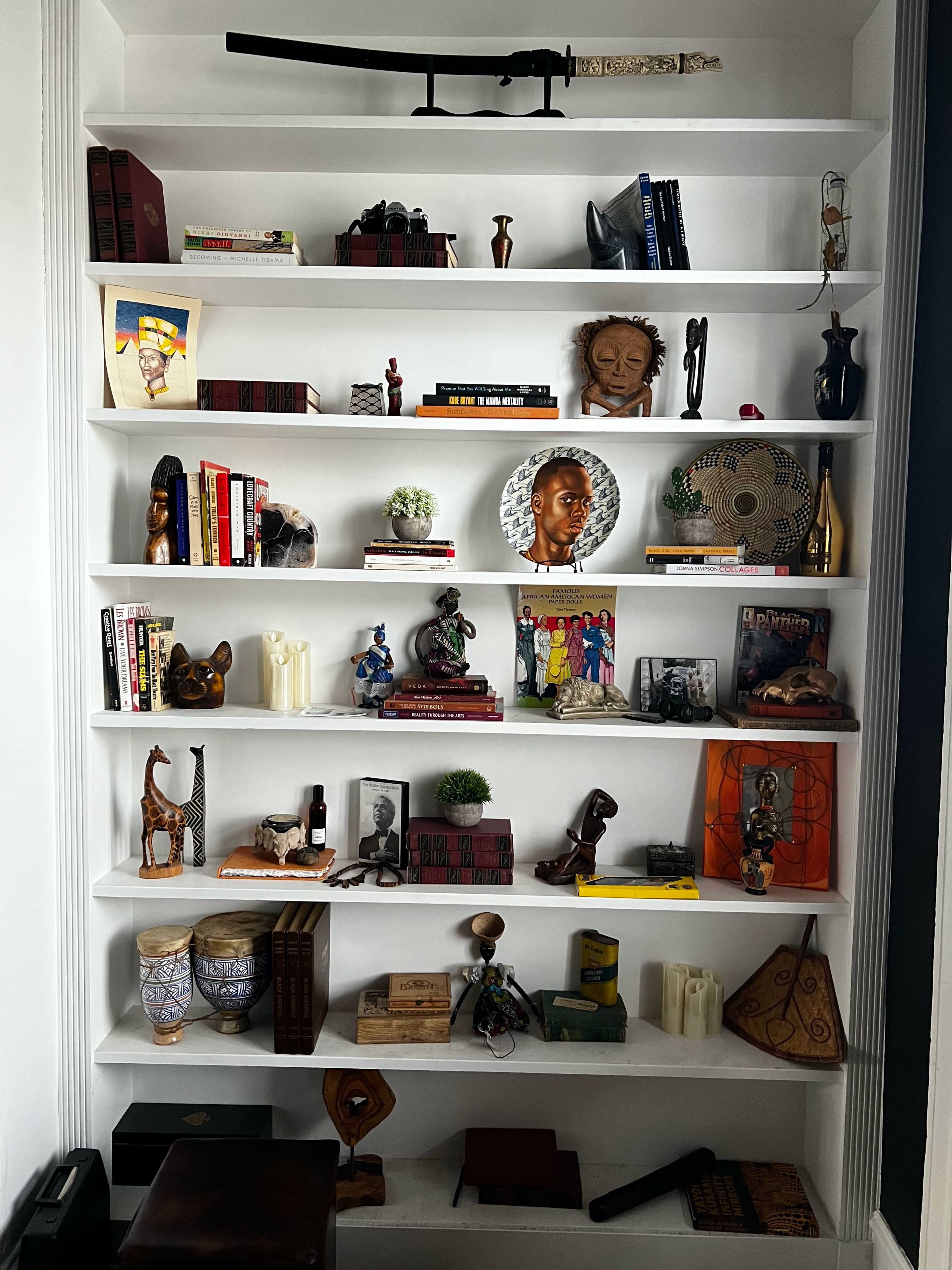 A white shelving unit filled with various decorative items, including books, sculptures, masks, and photographs arranged across several shelves.