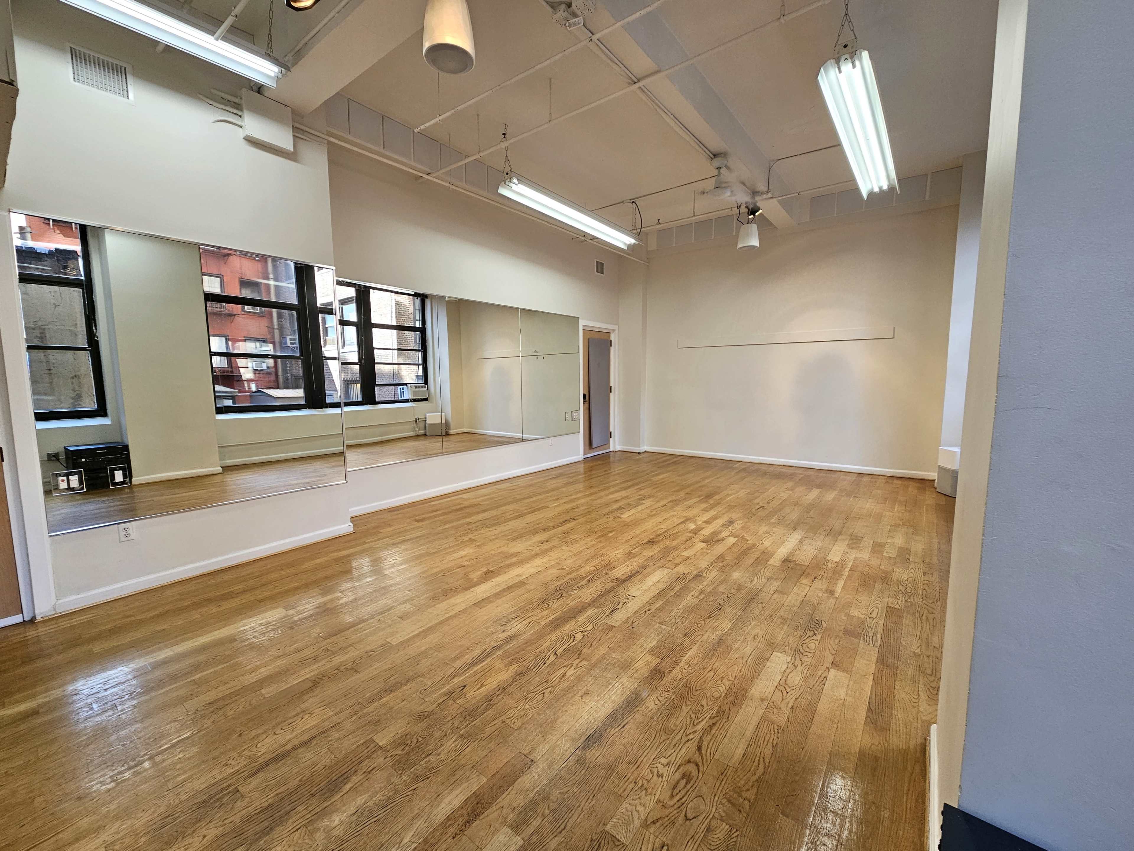 The image shows a spacious, empty room with hardwood floors and large windows, featuring a mirrored wall on one side.