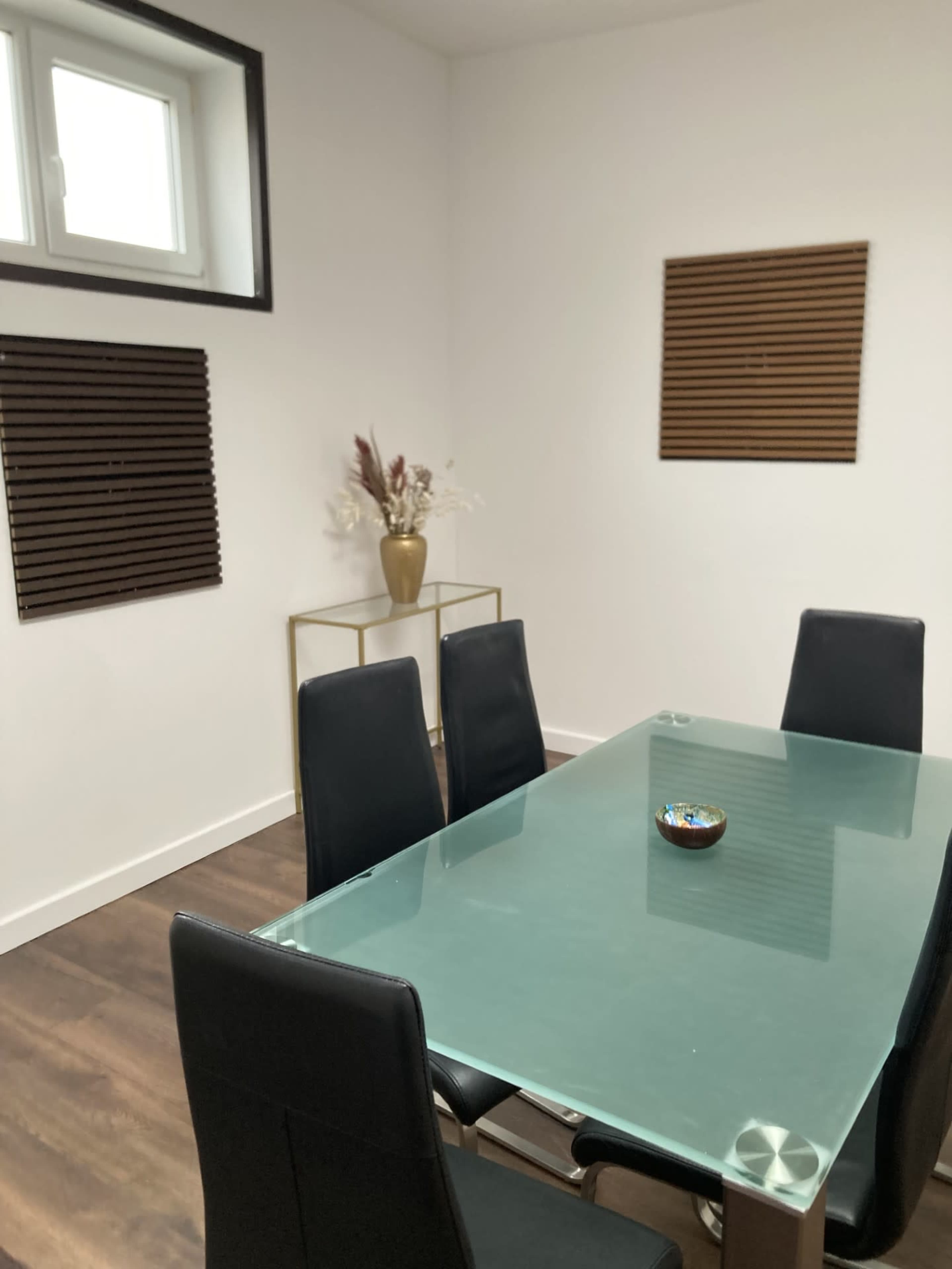 The image shows a modern dining area with a glass table, black chairs, and decorative wall panels, featuring a small vase on a gold-framed stand.