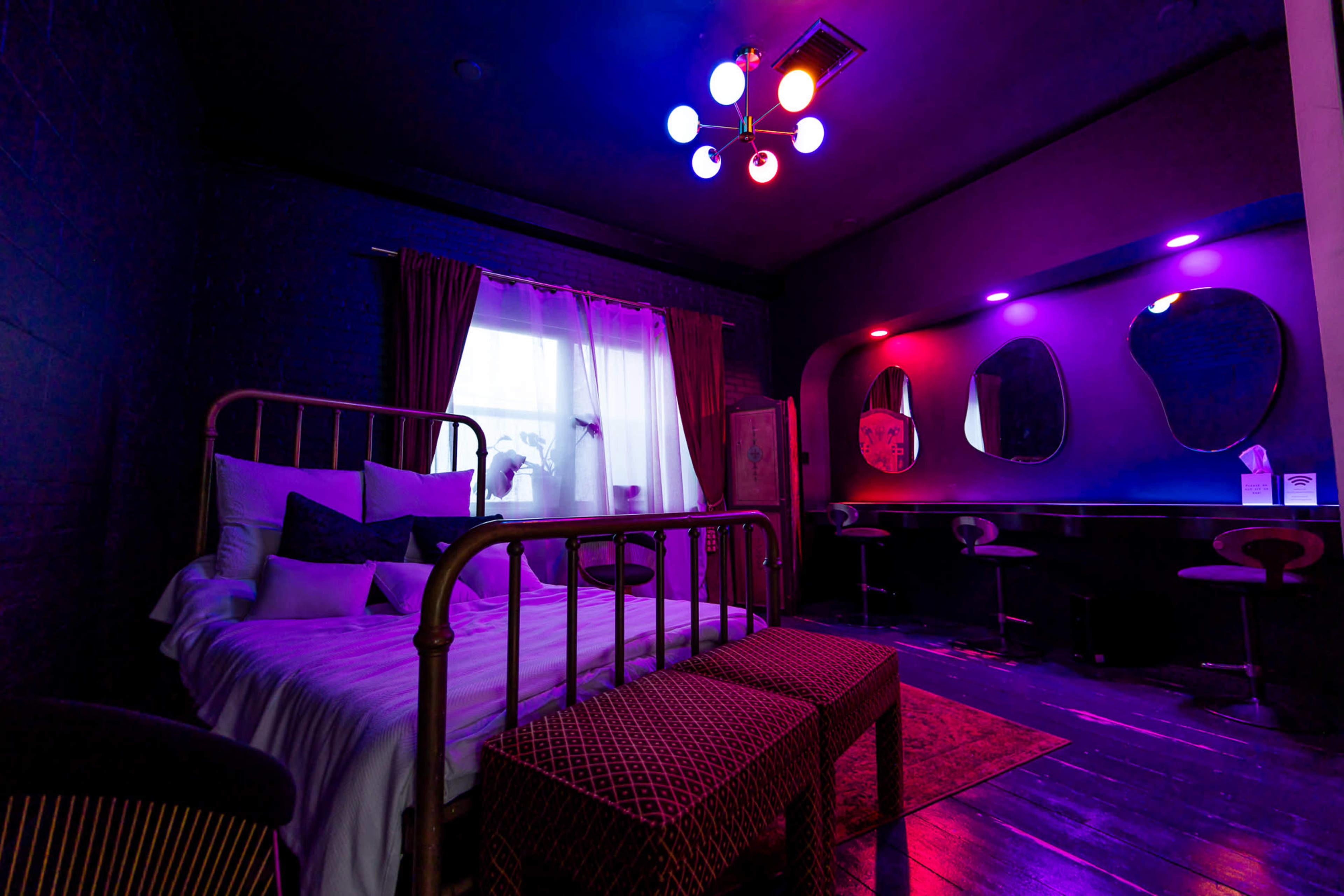 A bedroom features a metallic bed with a white bedspread, vibrant lighting, and a modern seating area with two mirrors.