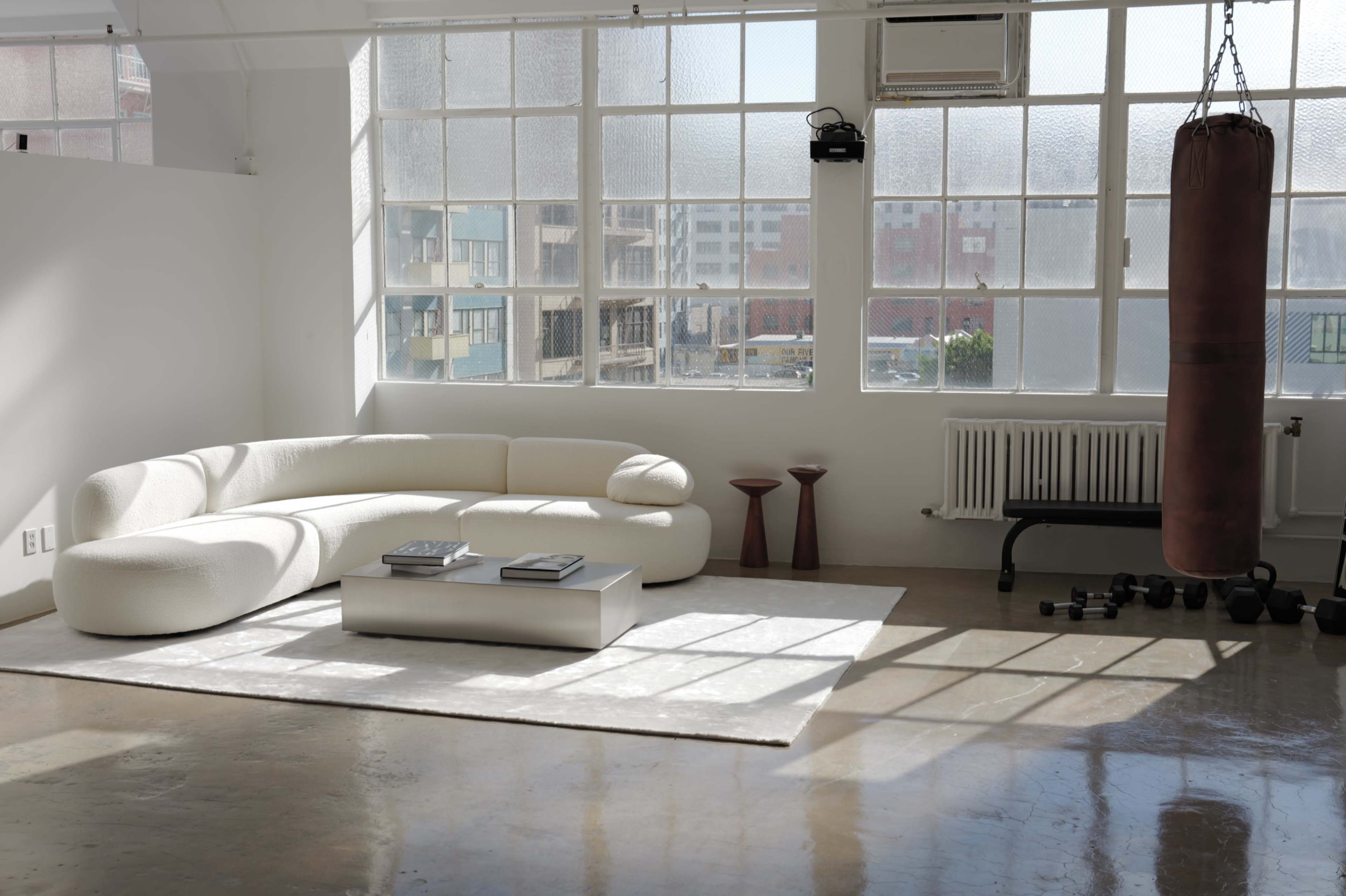 The image shows a bright and airy room featuring a curved white sofa, a coffee table with books, two small red stools, a punching bag, and decorative exercise weights.
