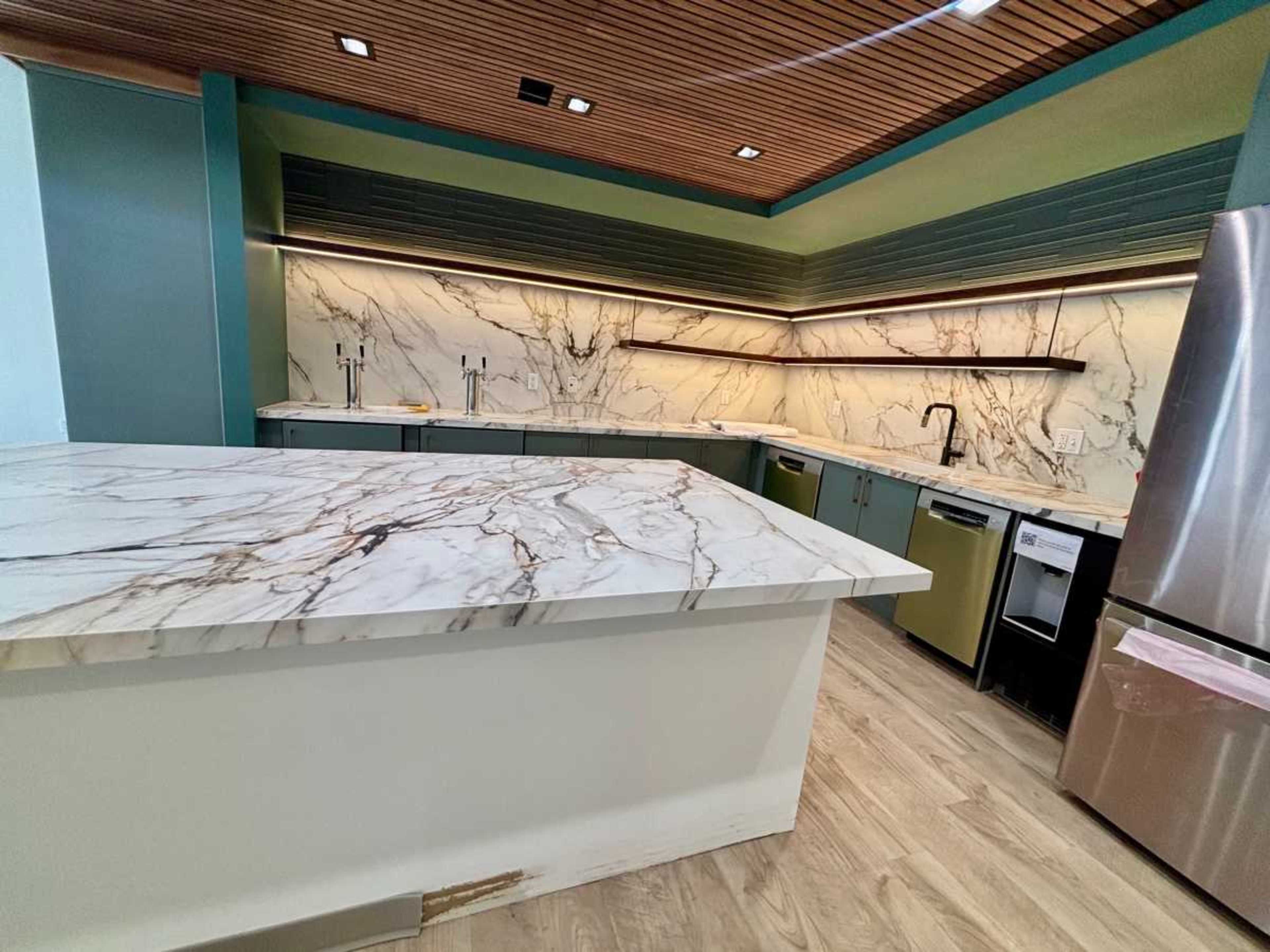 The image shows a modern kitchen featuring a large marble countertop, green cabinetry, and an accent wall with wooden paneling.