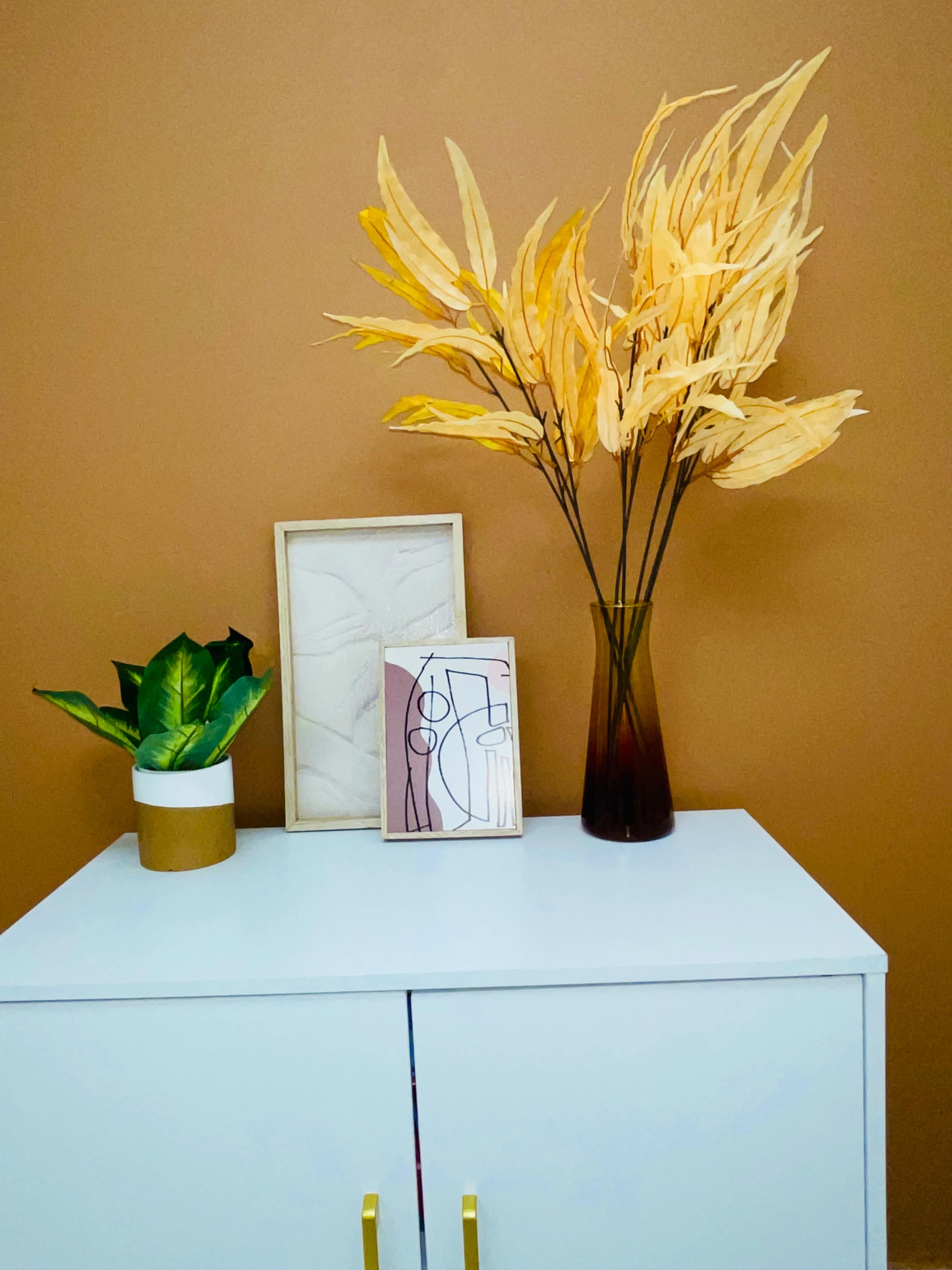 A white cabinet holds a green plant in a gold pot, a framed abstract artwork, and a vase of dried yellow leaves against a brown wall.