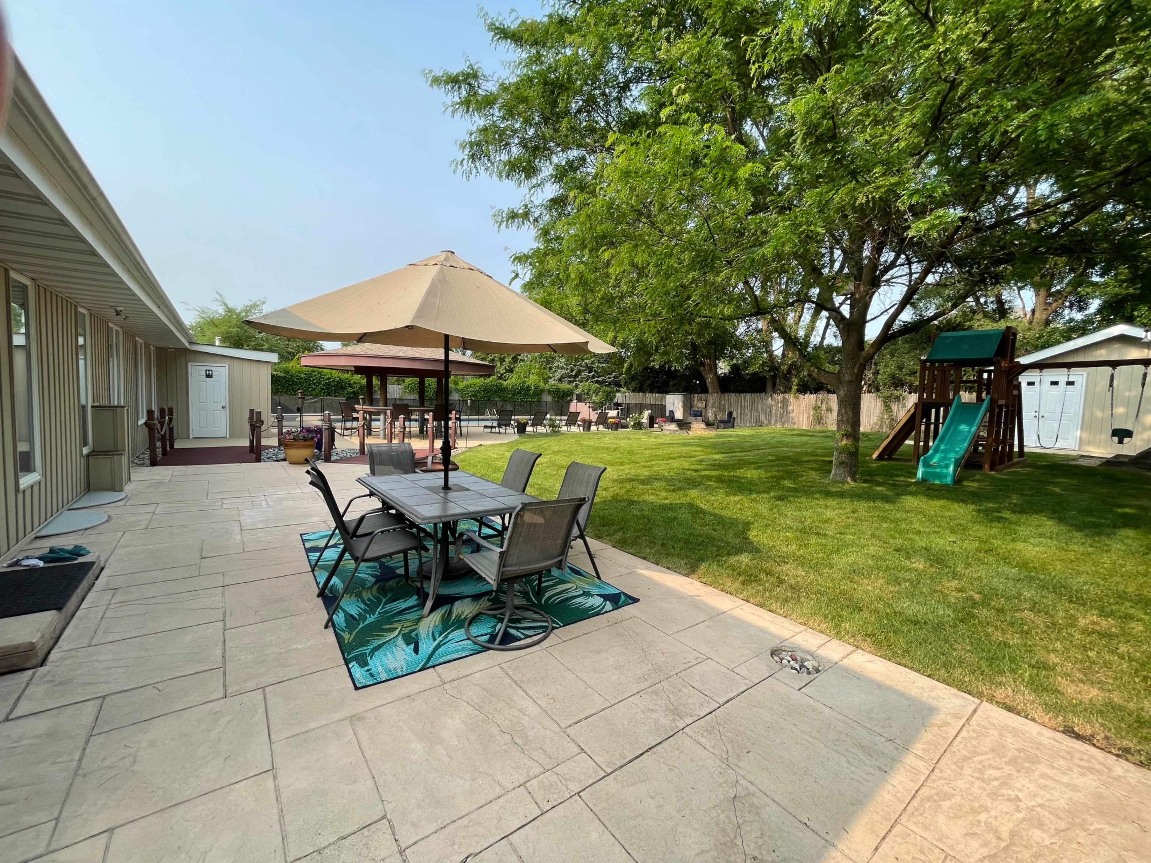 A backyard patio features a dining table under an umbrella, surrounded by grass and trees, with a playset and a shed in the background.
