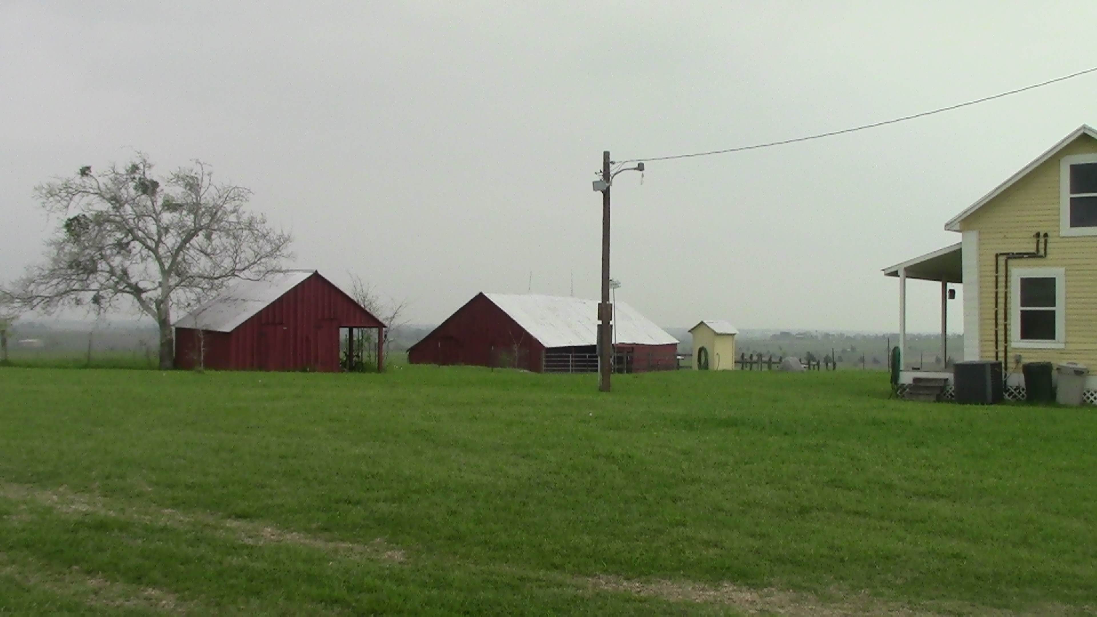 The image shows a rural landscape featuring several red barns, a yellow house, and a wide expanse of green grass under a cloudy sky.