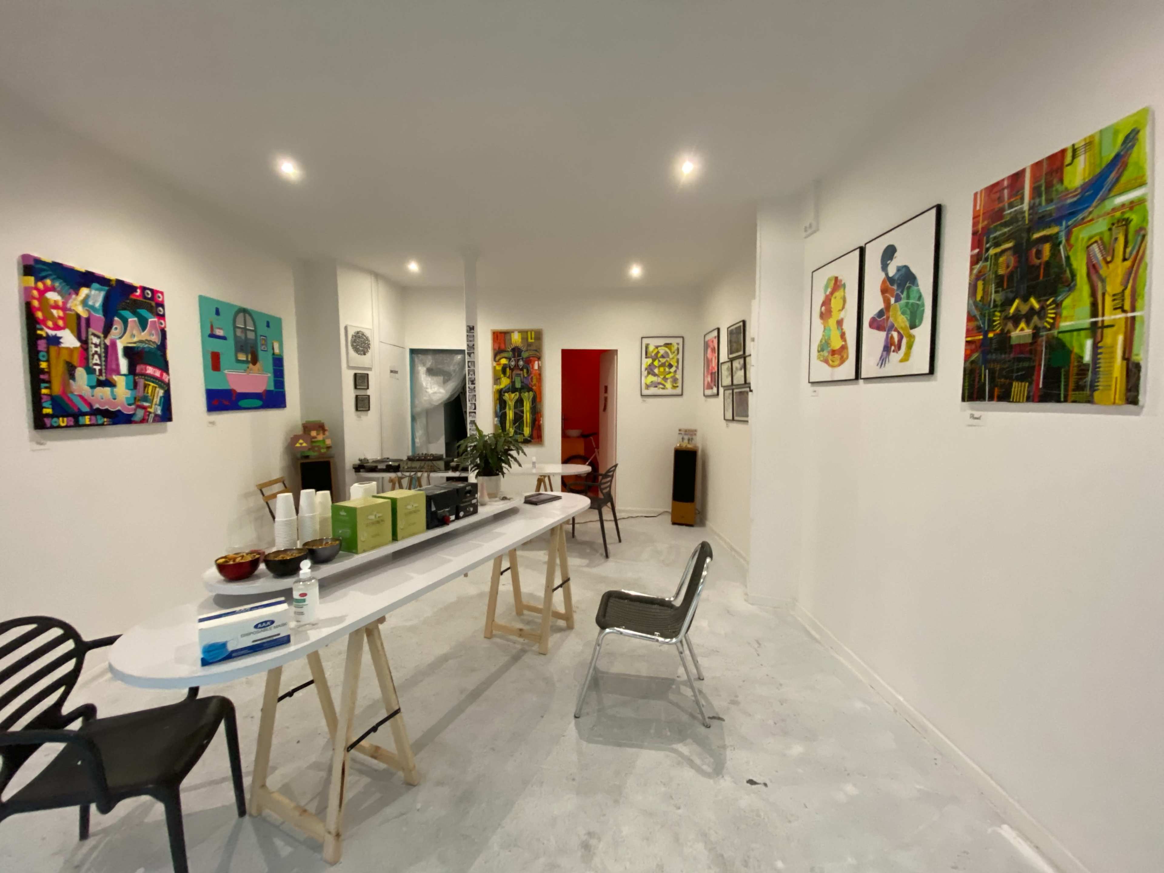 The image shows an art gallery featuring colorful paintings on the walls, with a table set up in the center displaying refreshments.