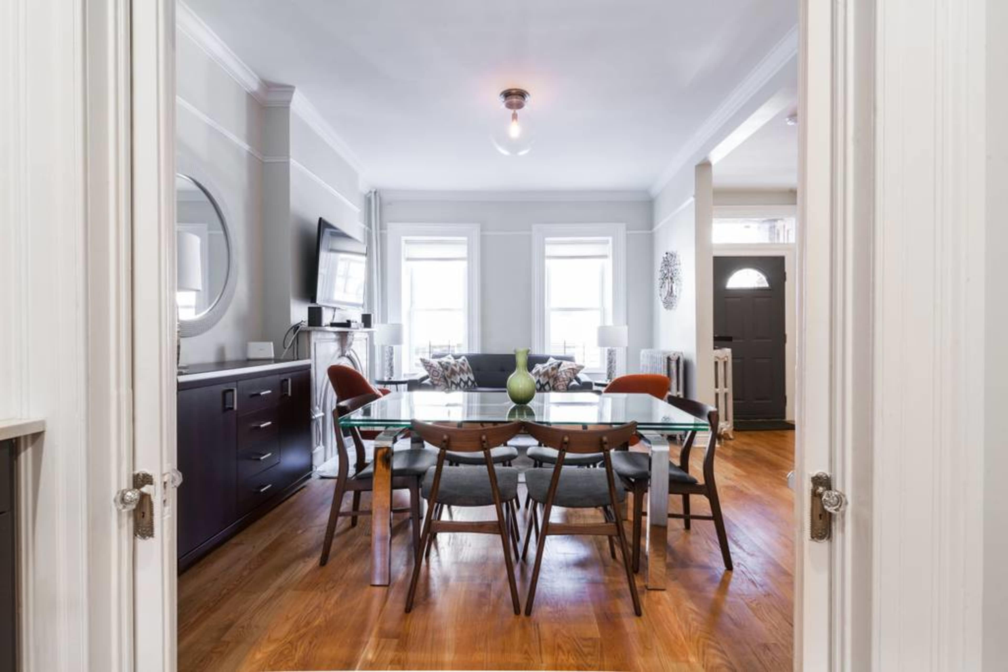 The image shows a dining area with a glass table surrounded by wooden chairs, viewed from a hallway leading to a living space with a couch and television in the background.