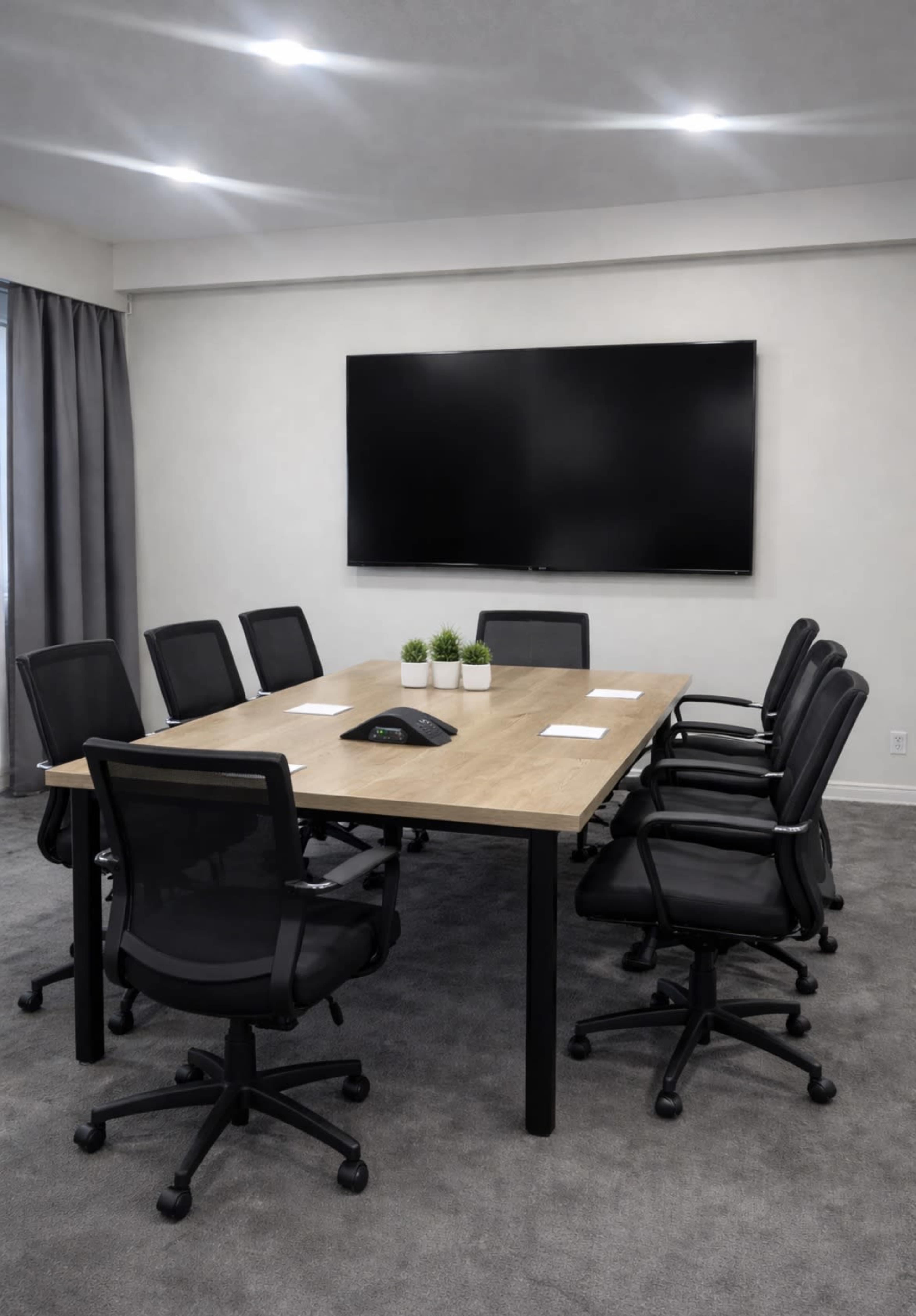 A conference room featuring a long wooden table surrounded by eight black office chairs, with a large screen mounted on the wall and small plants on the table.