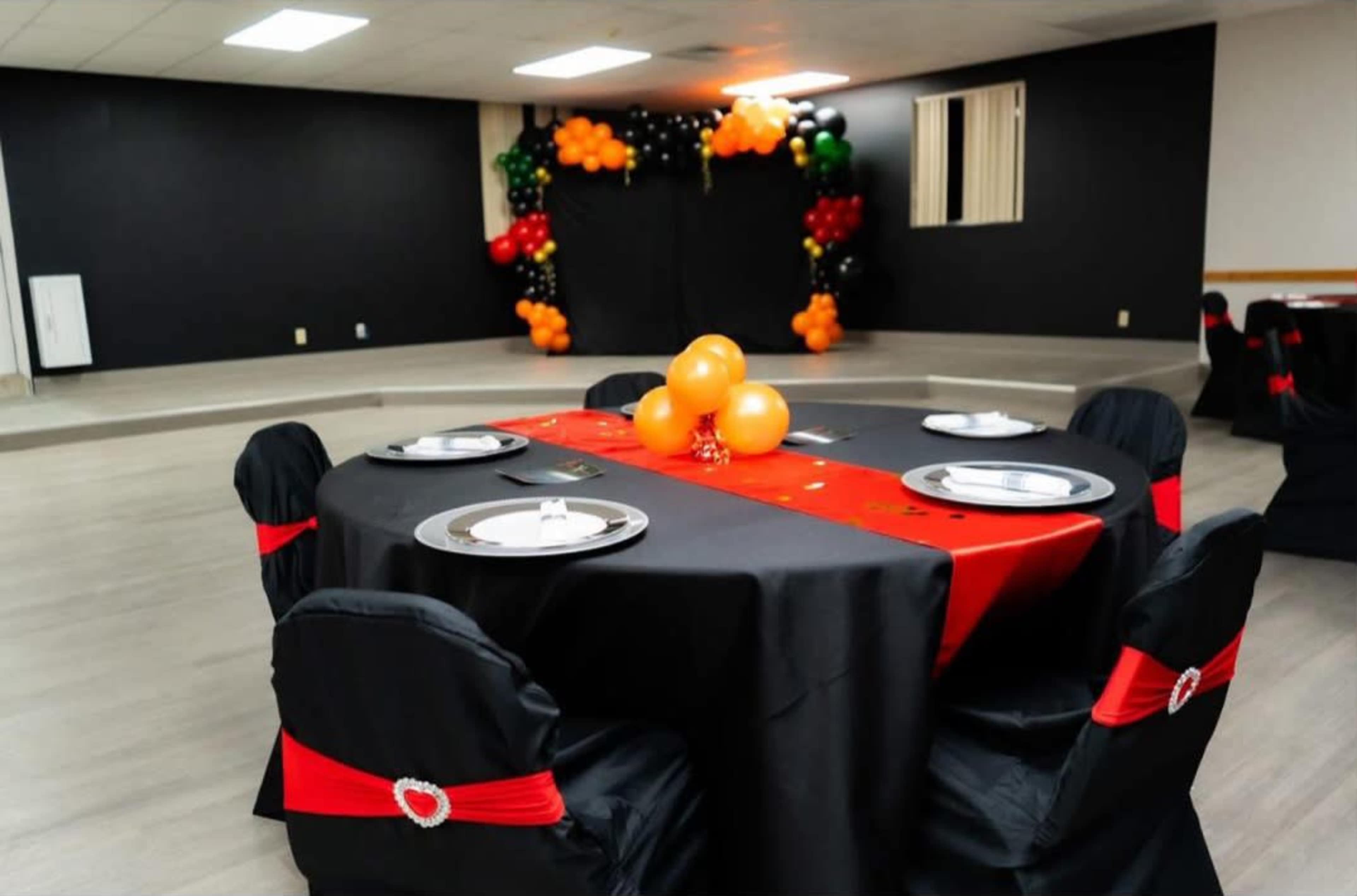 A decorated event space features a round table with a black and red tablecloth surrounded by chairs, and a balloon arch in the background.