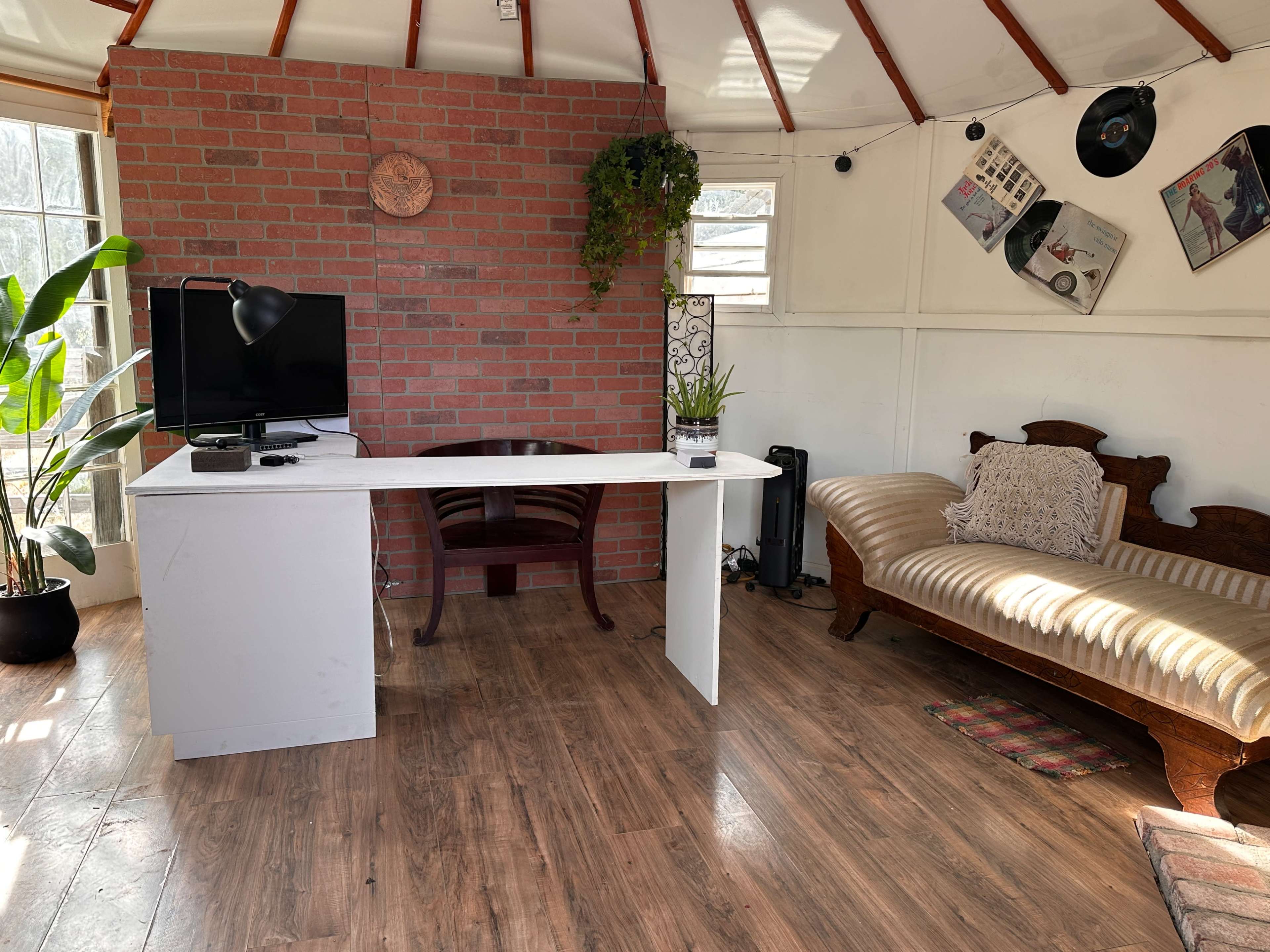 The image shows a cozy interior featuring a brick accent wall, a desk with a monitor, a chair, a plant, and a vintage sofa positioned in a circular room with wooden flooring.