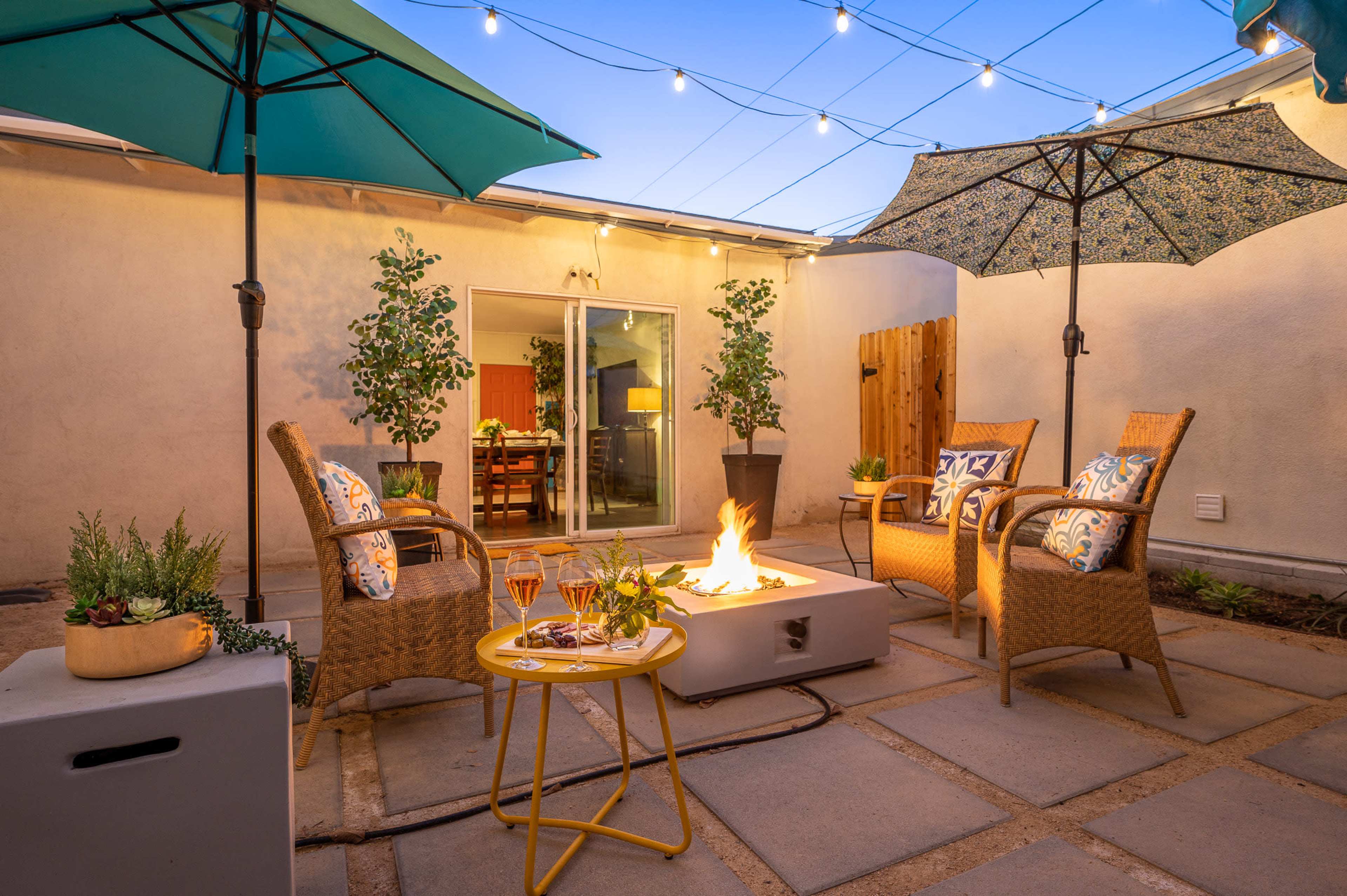 The image shows a cozy outdoor seating area with four chairs around a fire pit, surrounded by umbrellas and small plants, illuminated by string lights overhead.