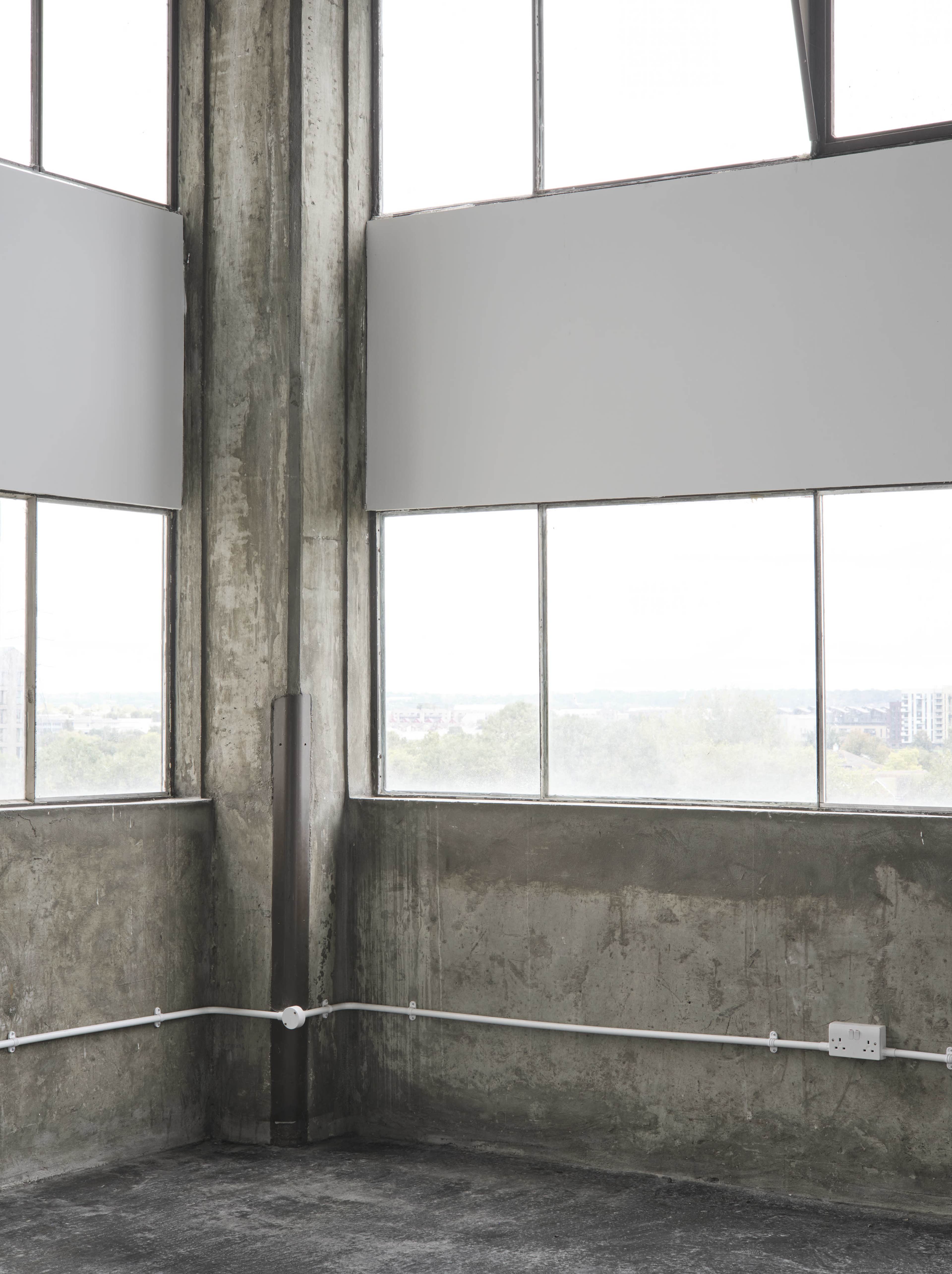 The image shows a corner of a room with large windows, concrete walls, and exposed plumbing.