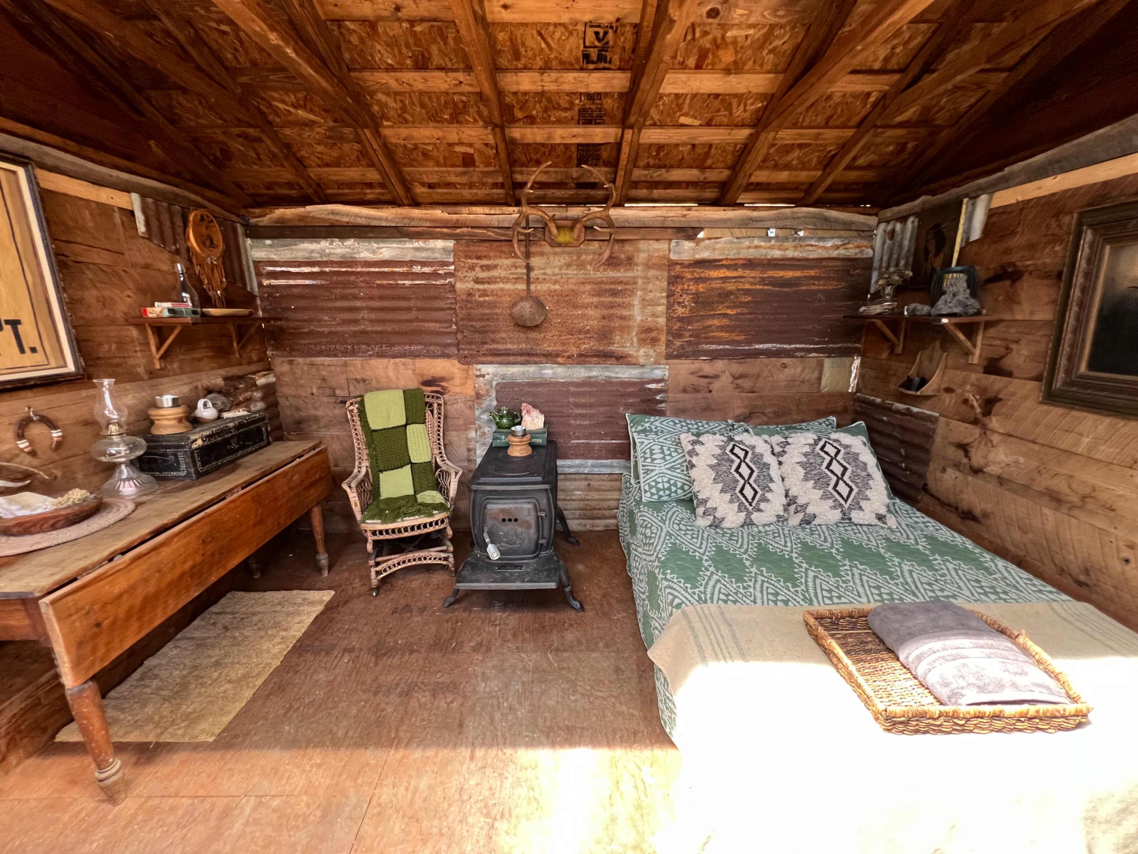 The image shows a rustic wooden cabin interior with a bed, a small stove, a table, and various decorative items on the walls.