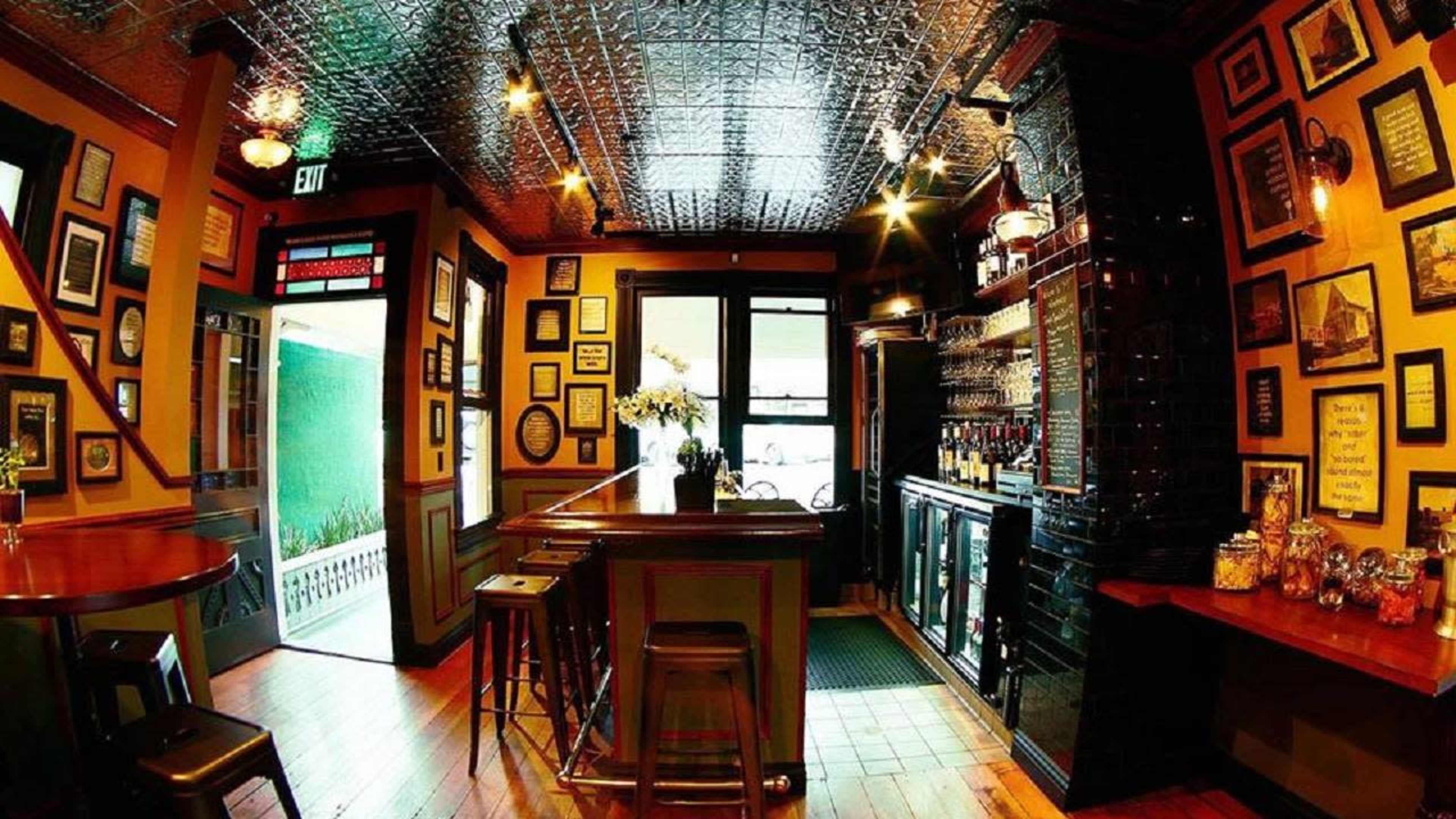 The image shows a cozy bar with wooden floors, a long counter, and walls adorned with framed pictures and decorations.