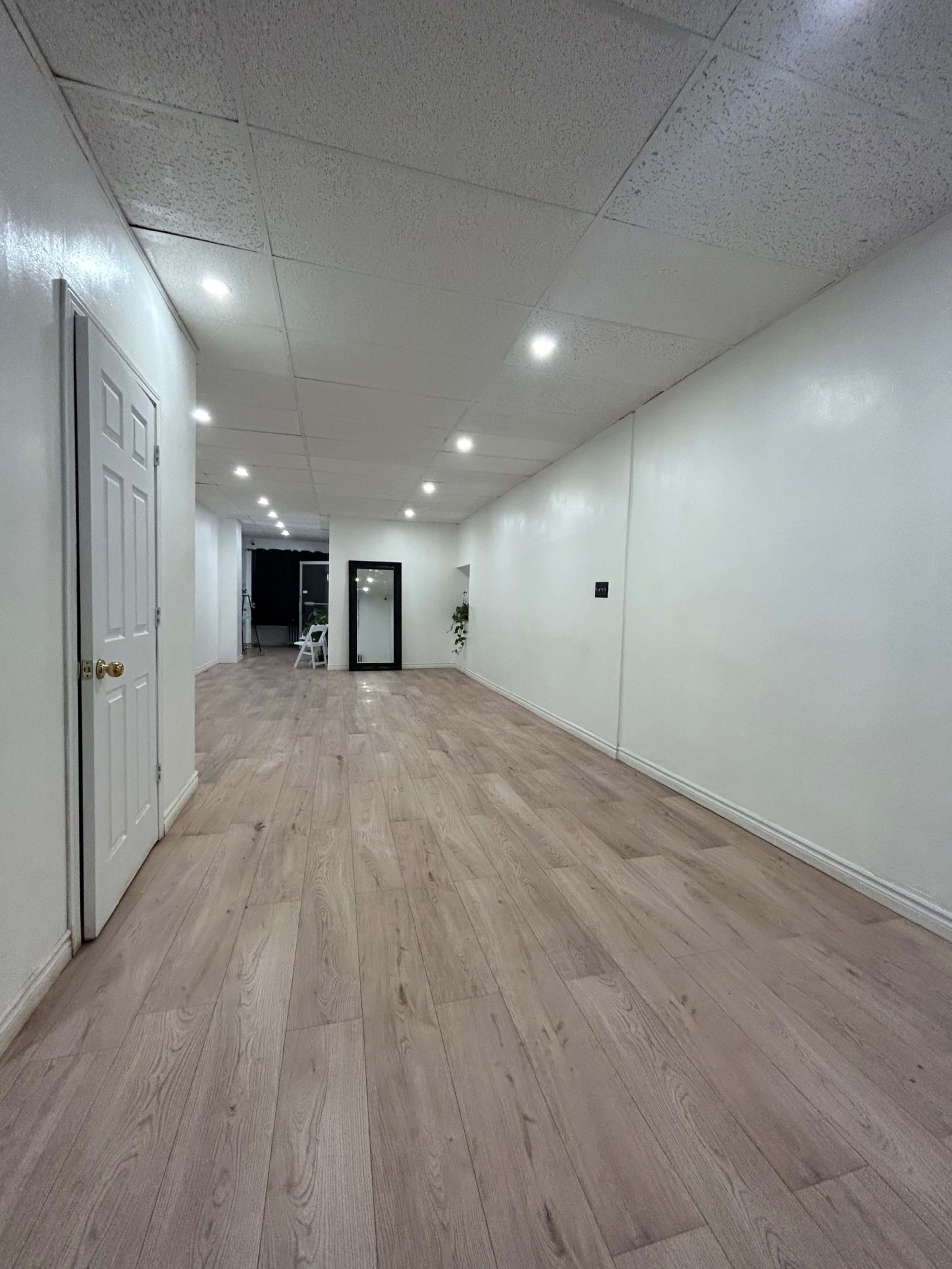 A long, empty hallway with white walls, wooden flooring, and lit by ceiling lights.
