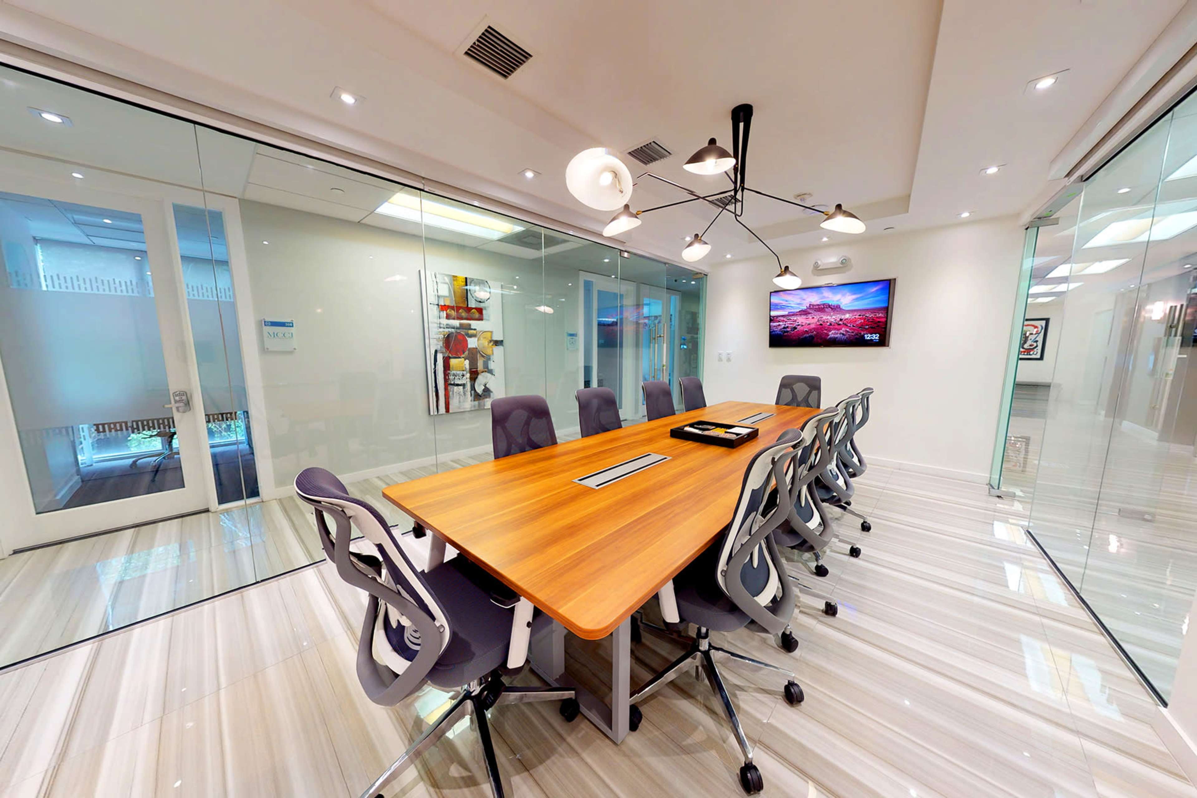 A modern conference room features a long wooden table surrounded by ergonomic chairs, with glass walls and a large artwork on display.