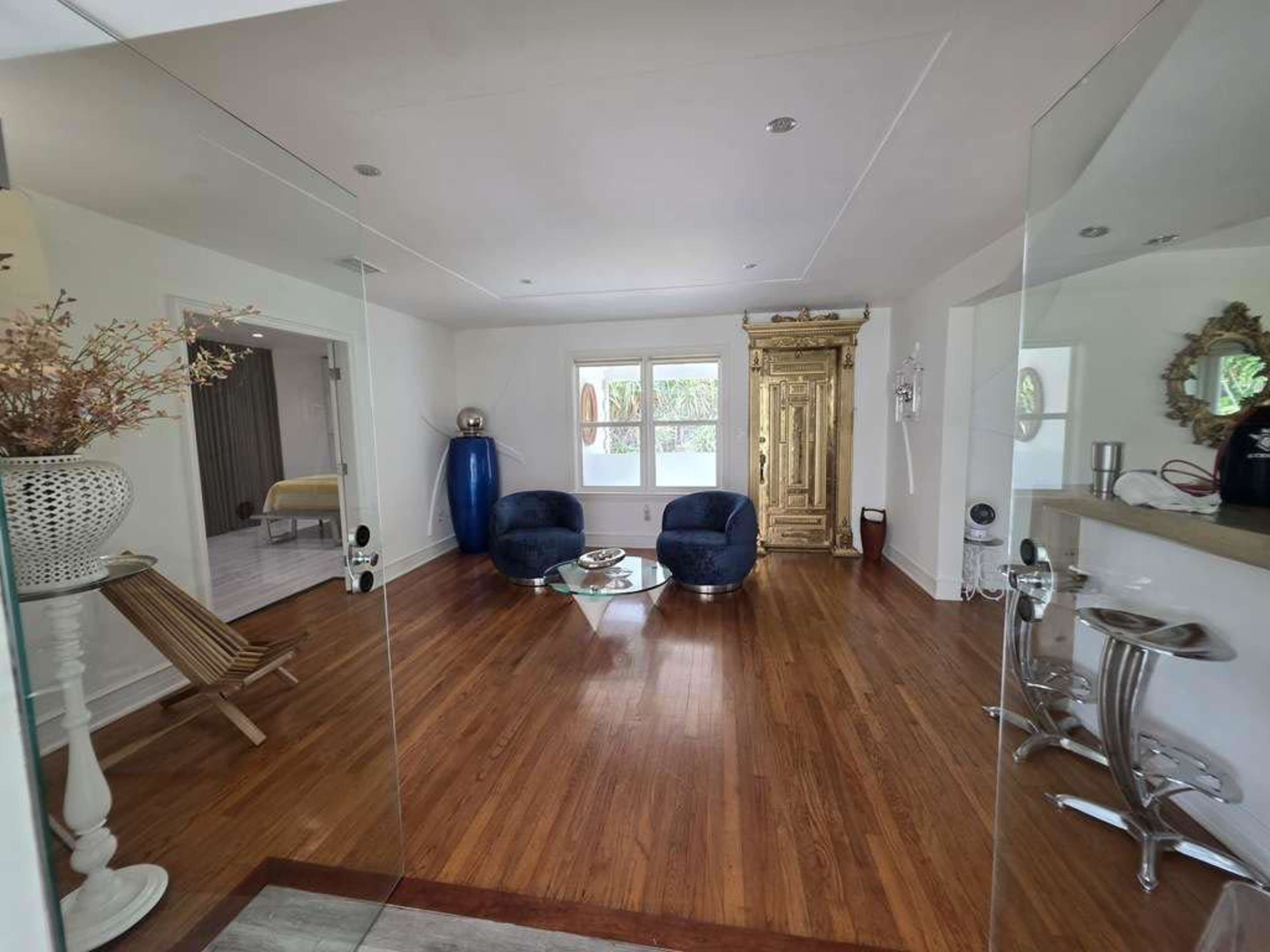 The image shows a modern living room with wooden floors, a glass partition, two blue armchairs, and a decorative golden cabinet.