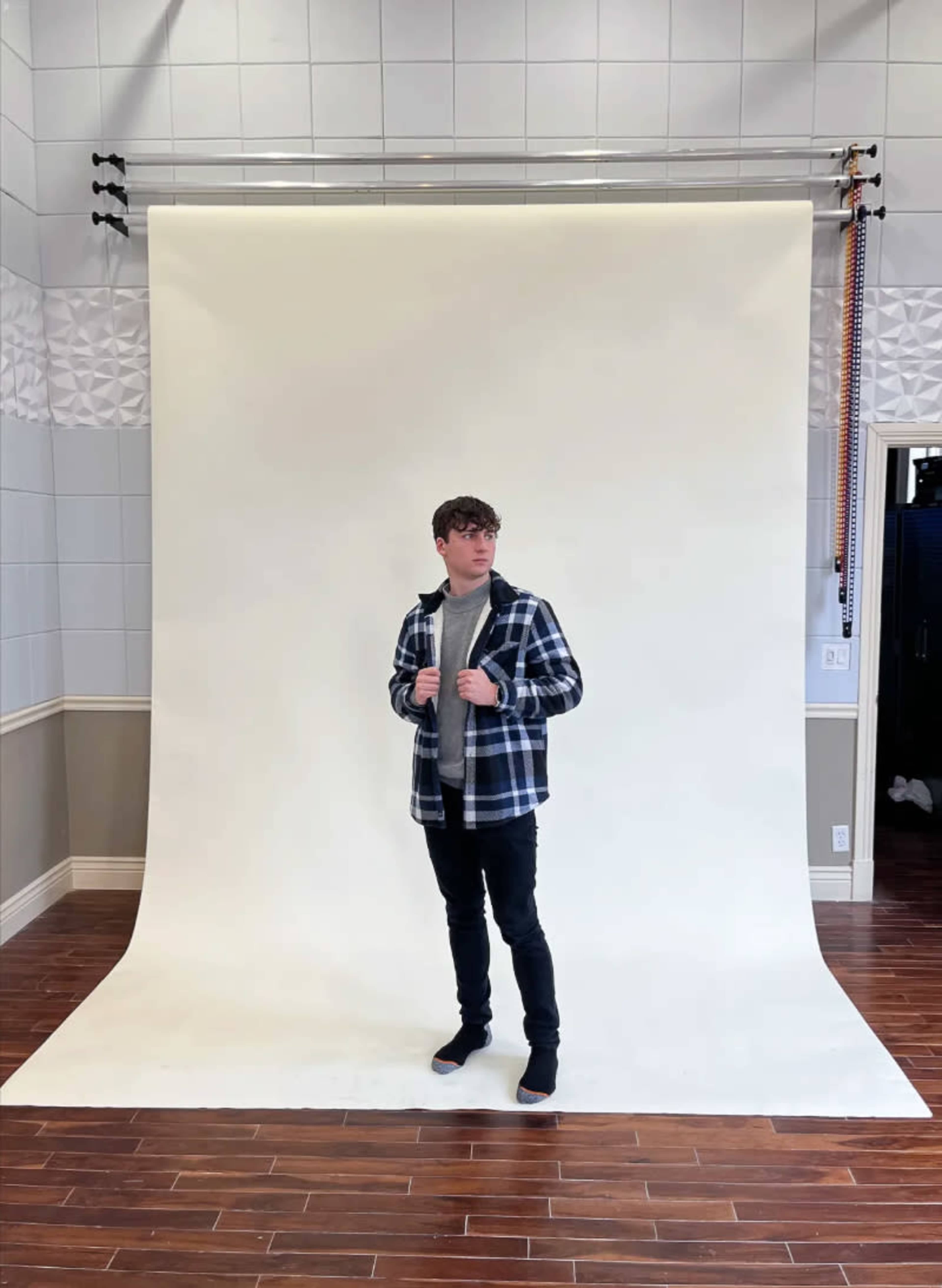 A person stands on a light-colored backdrop in a spacious studio with wooden flooring and a patterned wall.