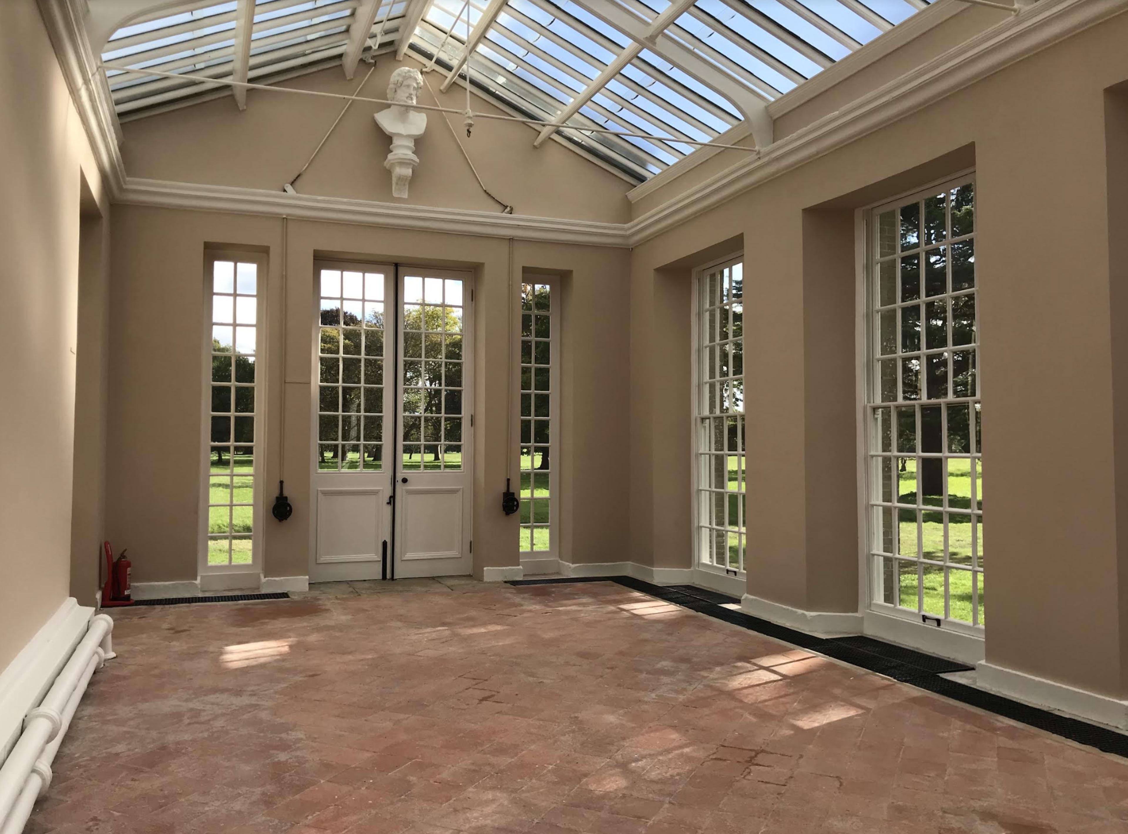 A sunlit room with large glass windows, a door in the center, and a terracotta tiled floor.