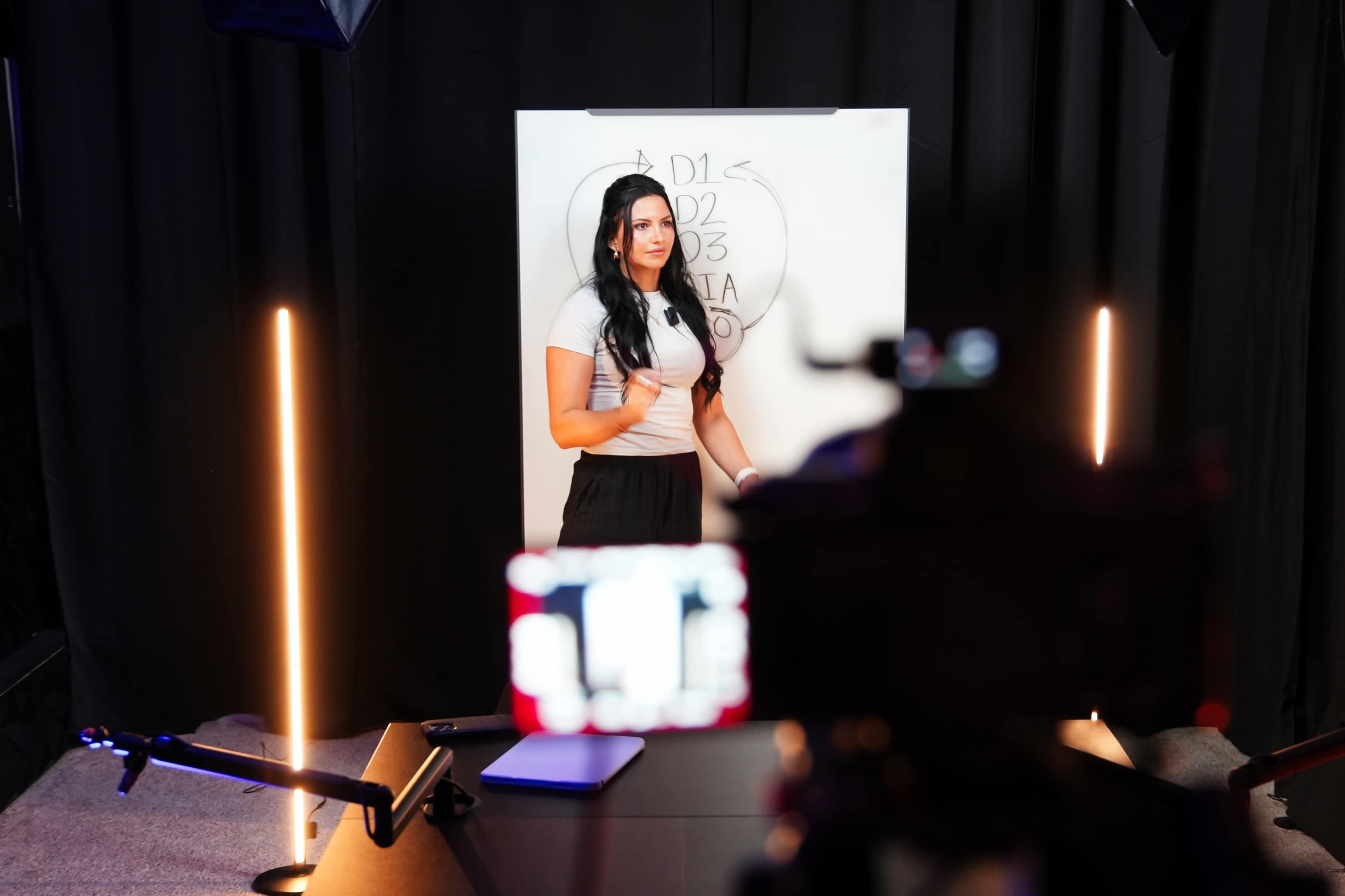 A woman stands in front of a whiteboard with a diagram, while a camera captures the scene from a nearby desk.