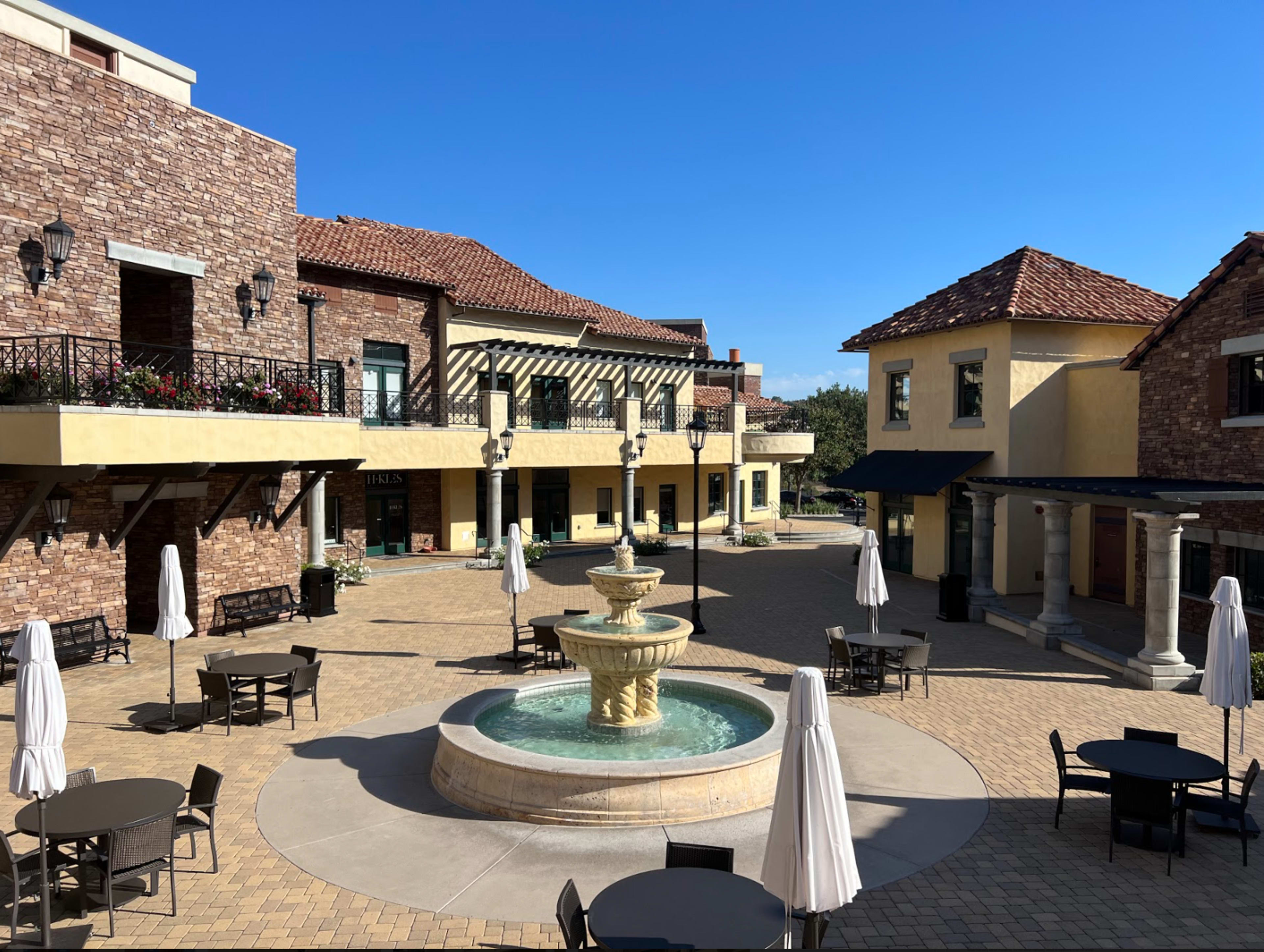 A courtyard with a fountain surrounded by several buildings featuring a mix of architectural styles, along with patio tables and umbrellas.