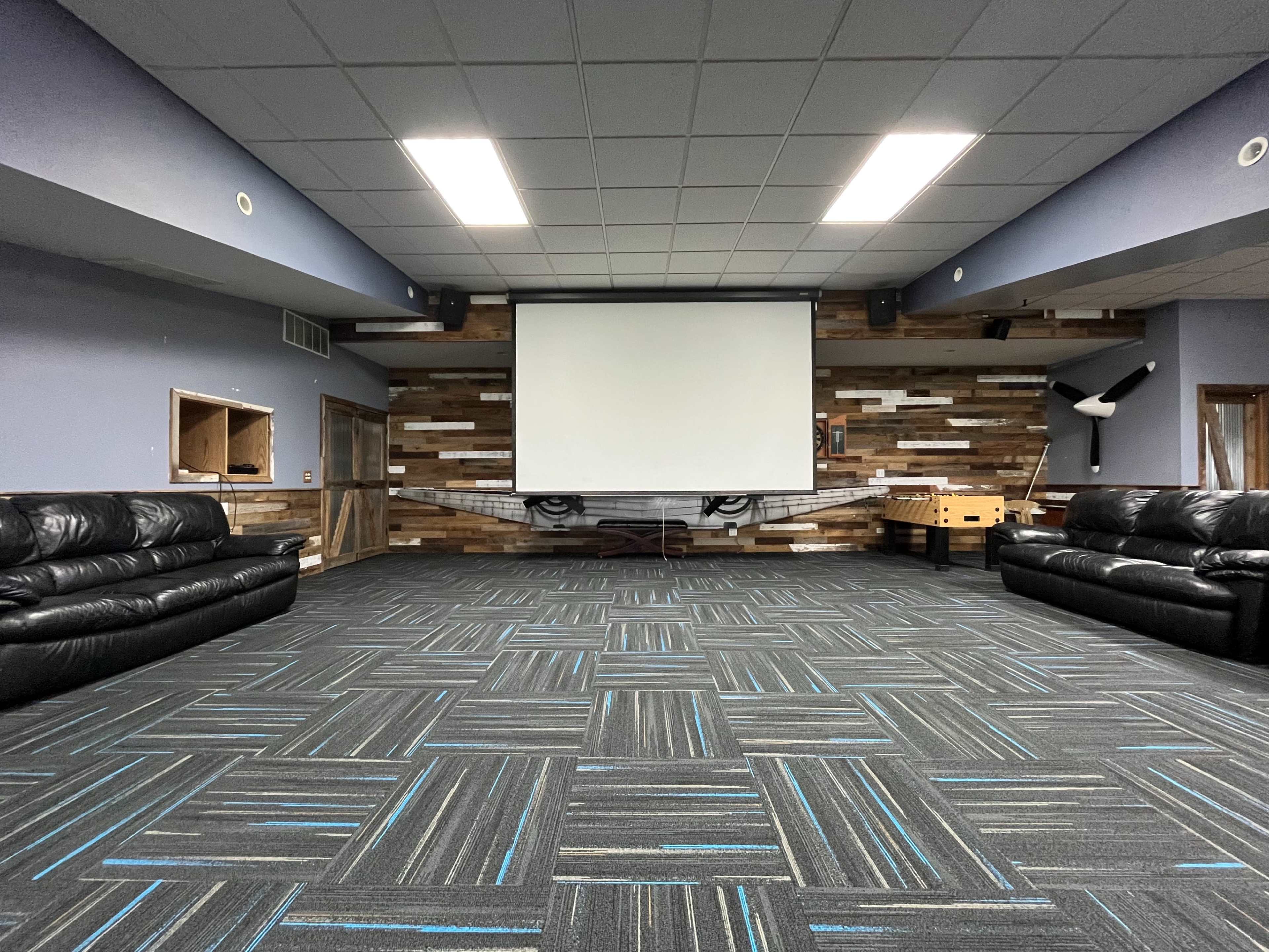The image shows a spacious room with two black leather couches facing a blank projection screen against a wooden feature wall.
