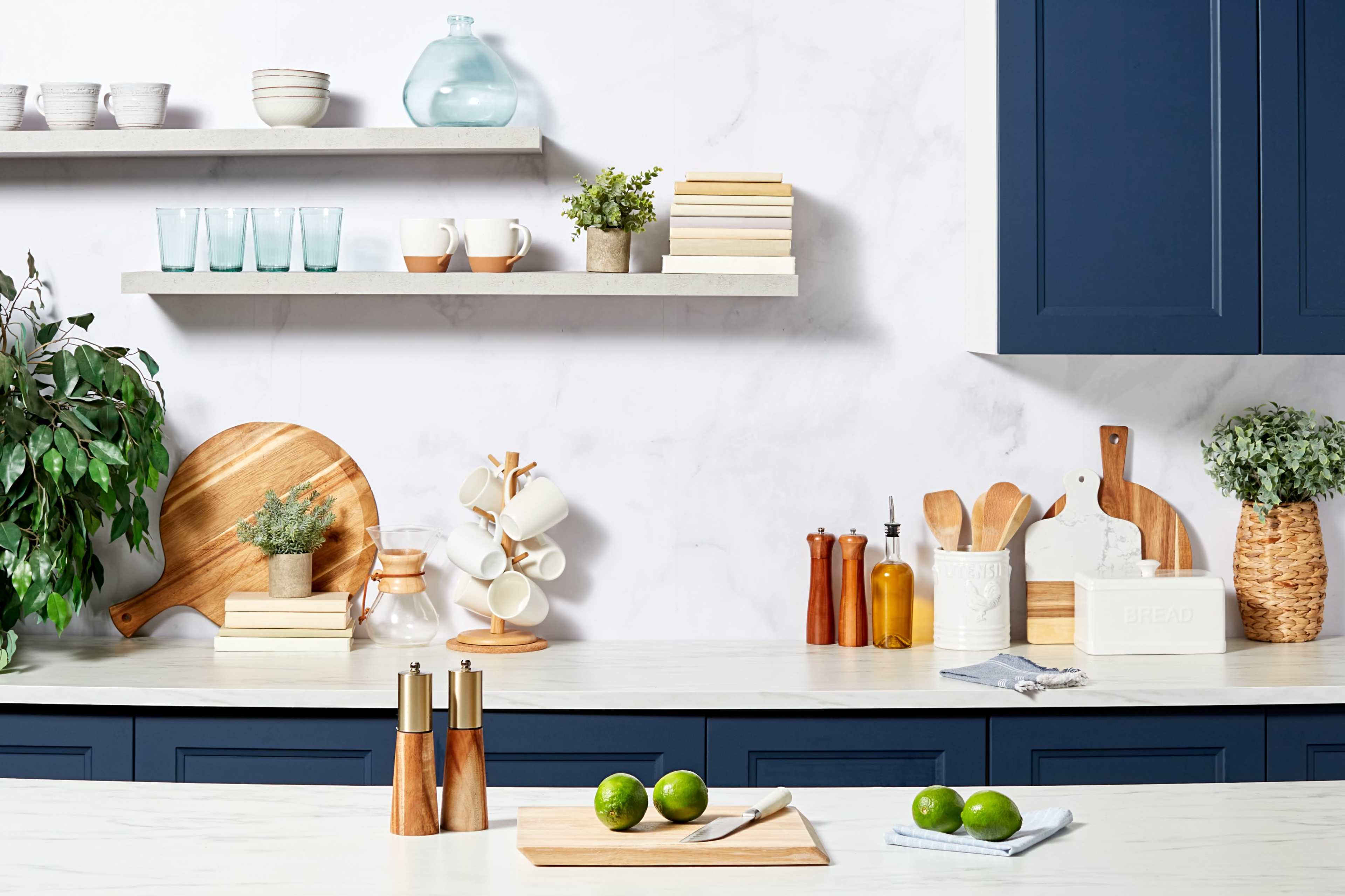 A modern kitchen countertop features a cutting board with limes, wooden utensils, glass containers, and decorative plants against a backdrop of navy cabinetry and floating shelves.