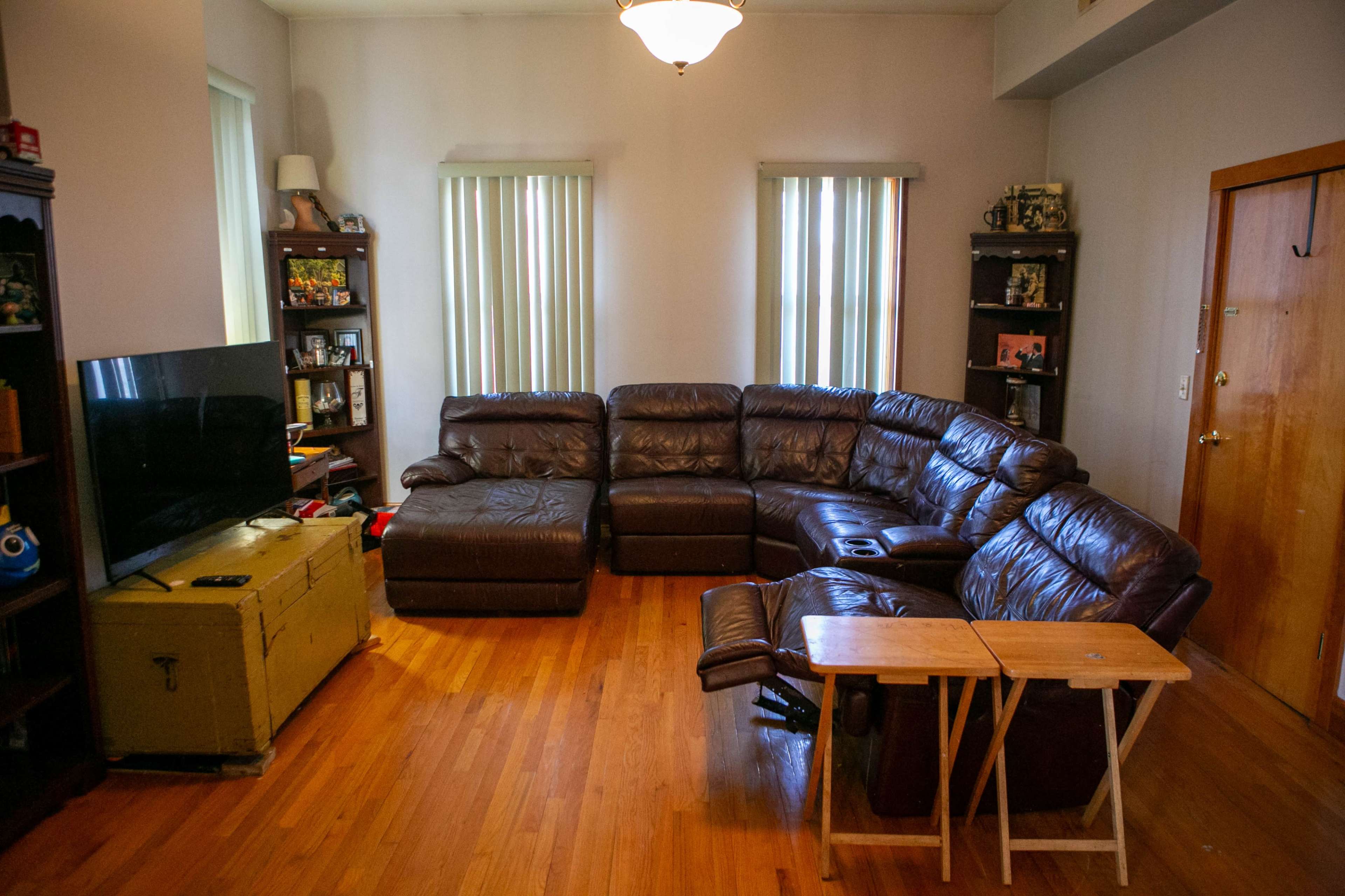 The image shows a living room with a brown sectional couch, a flat-screen TV on a wooden stand, and a table next to the couch, all set against wooden flooring and window treatments.