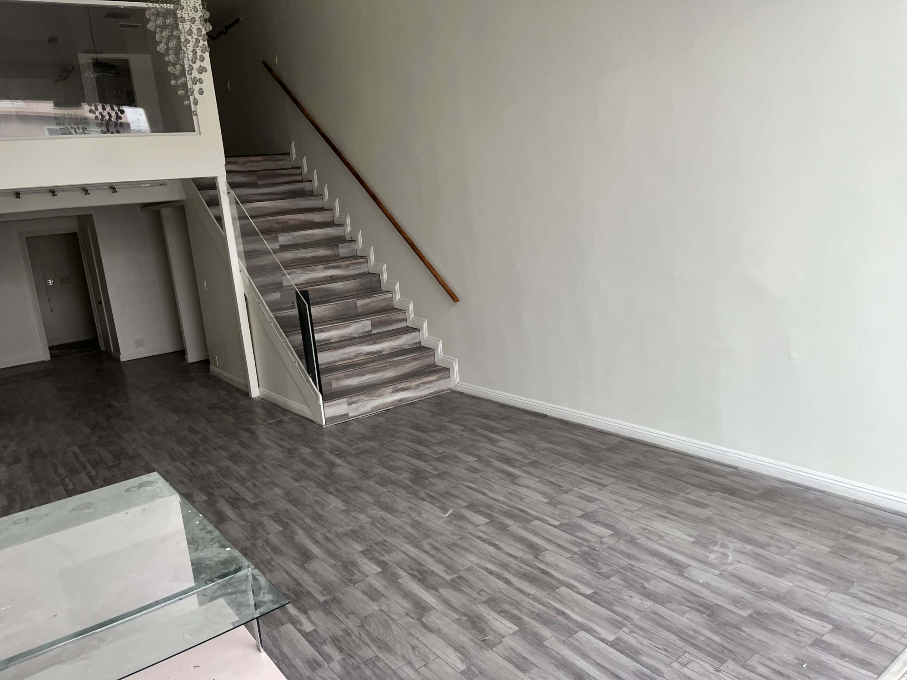 The image shows a spacious, empty room with light-colored walls, a staircase along one side, and a wooden floor.