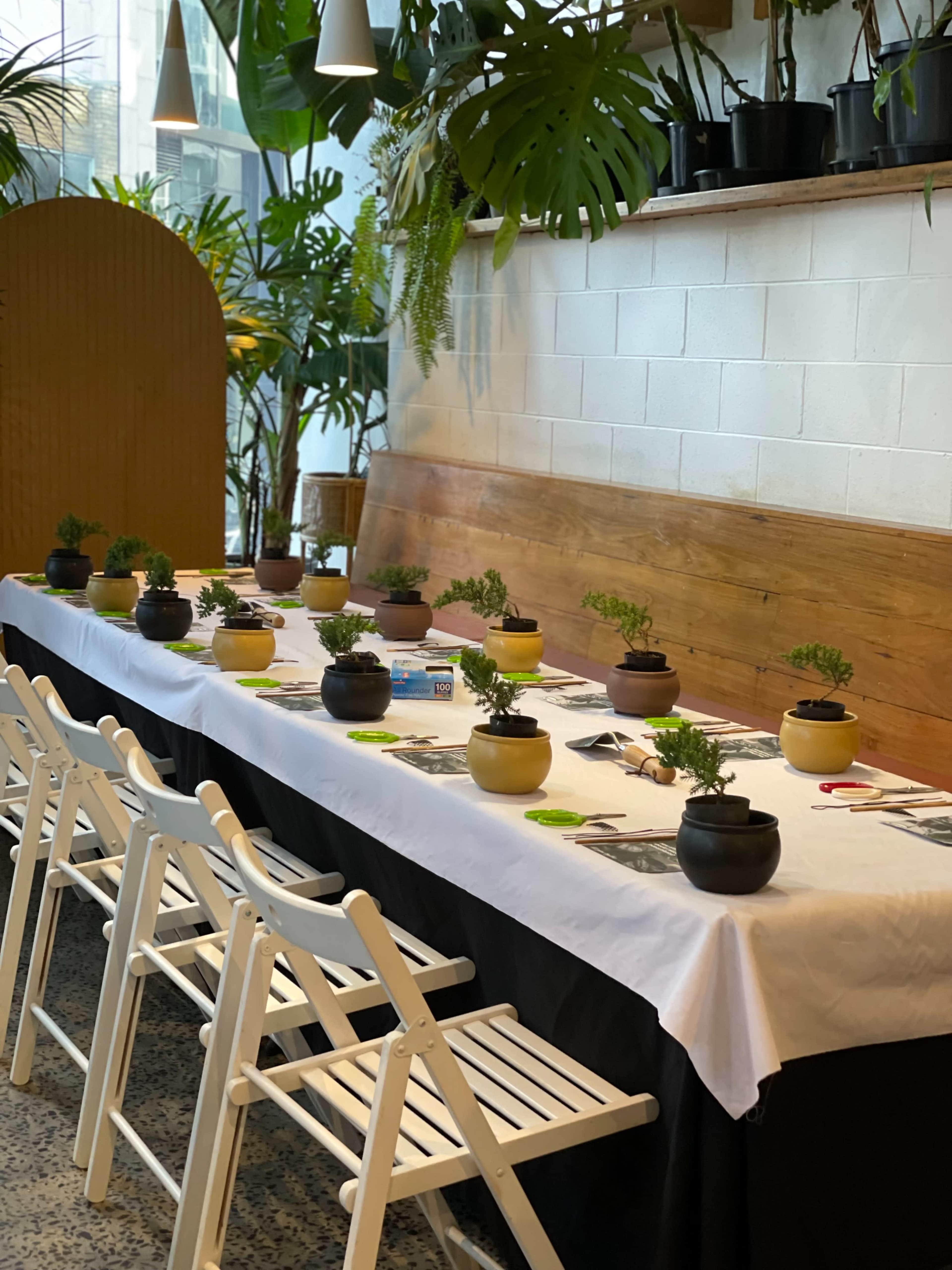A long table is set with white tablecloths and black and yellow planters containing small plants, surrounded by white folding chairs in a greenery-filled space.
